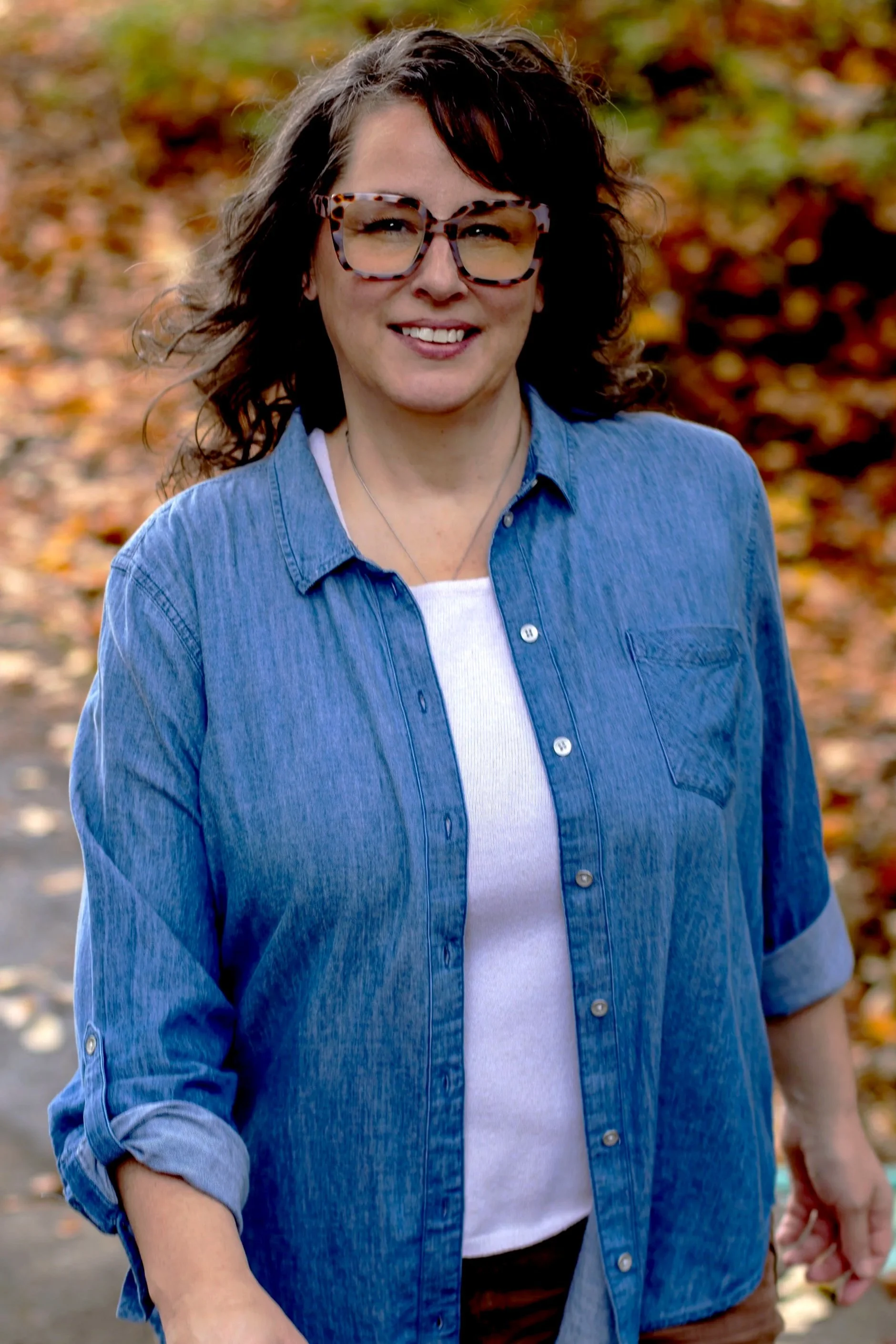 A woman with curly dark hair and glasses, wearing a denim shirt over a white t-shirt, smiling outdoors in front of autumn leaves.