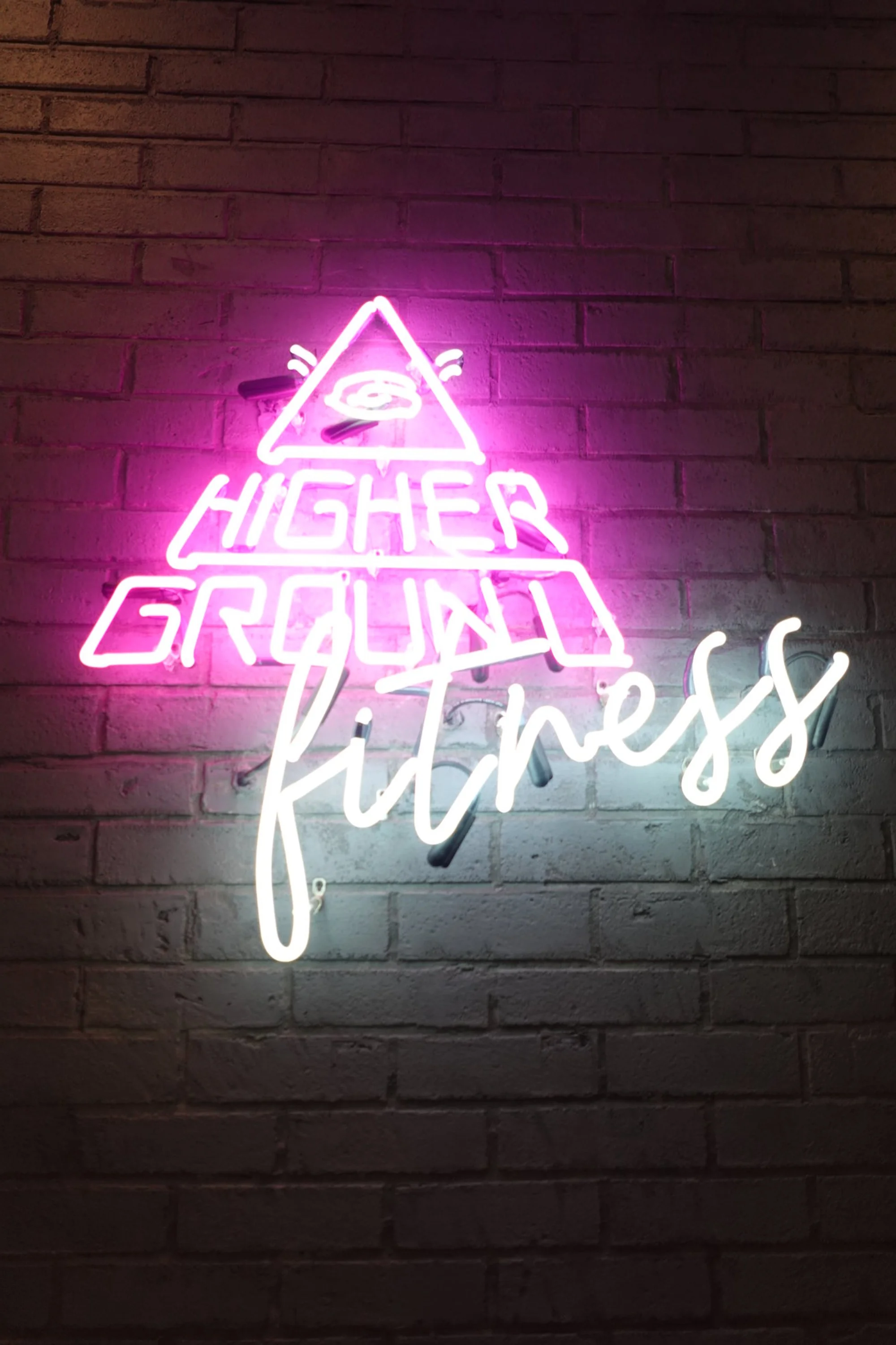 A neon sign that reads 'Higher Ground Fitness' with a triangle symbol and spiral design in pink and white, mounted on a brick wall.