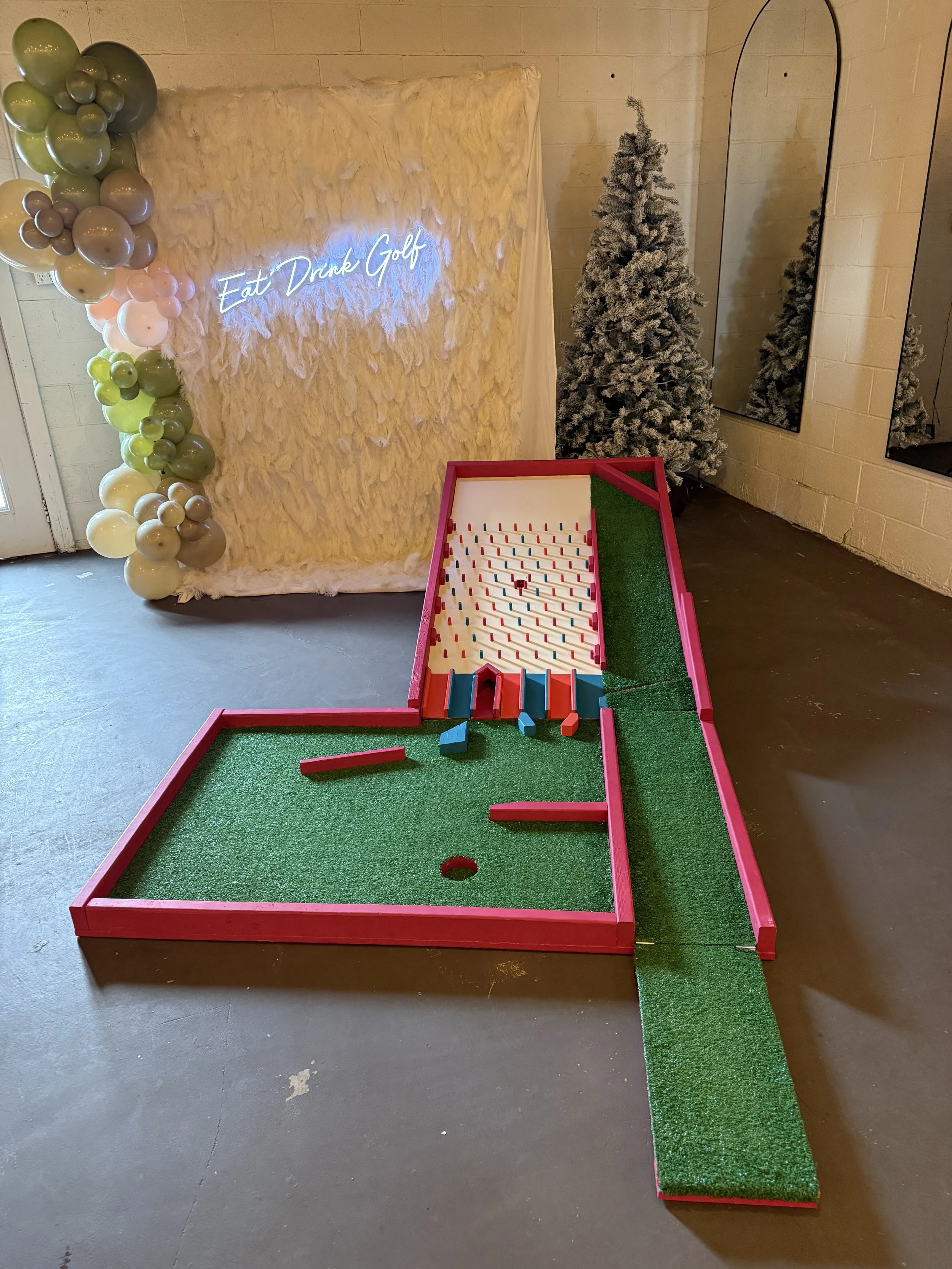 Miniature outdoor golf game with artificial turf, putting hole, and obstacles, surrounded by holiday decorations including a snow-dusted Christmas tree, balloons, and a fuzzy backdrop with neon lettering that reads 'Eat Drink Golf'.
