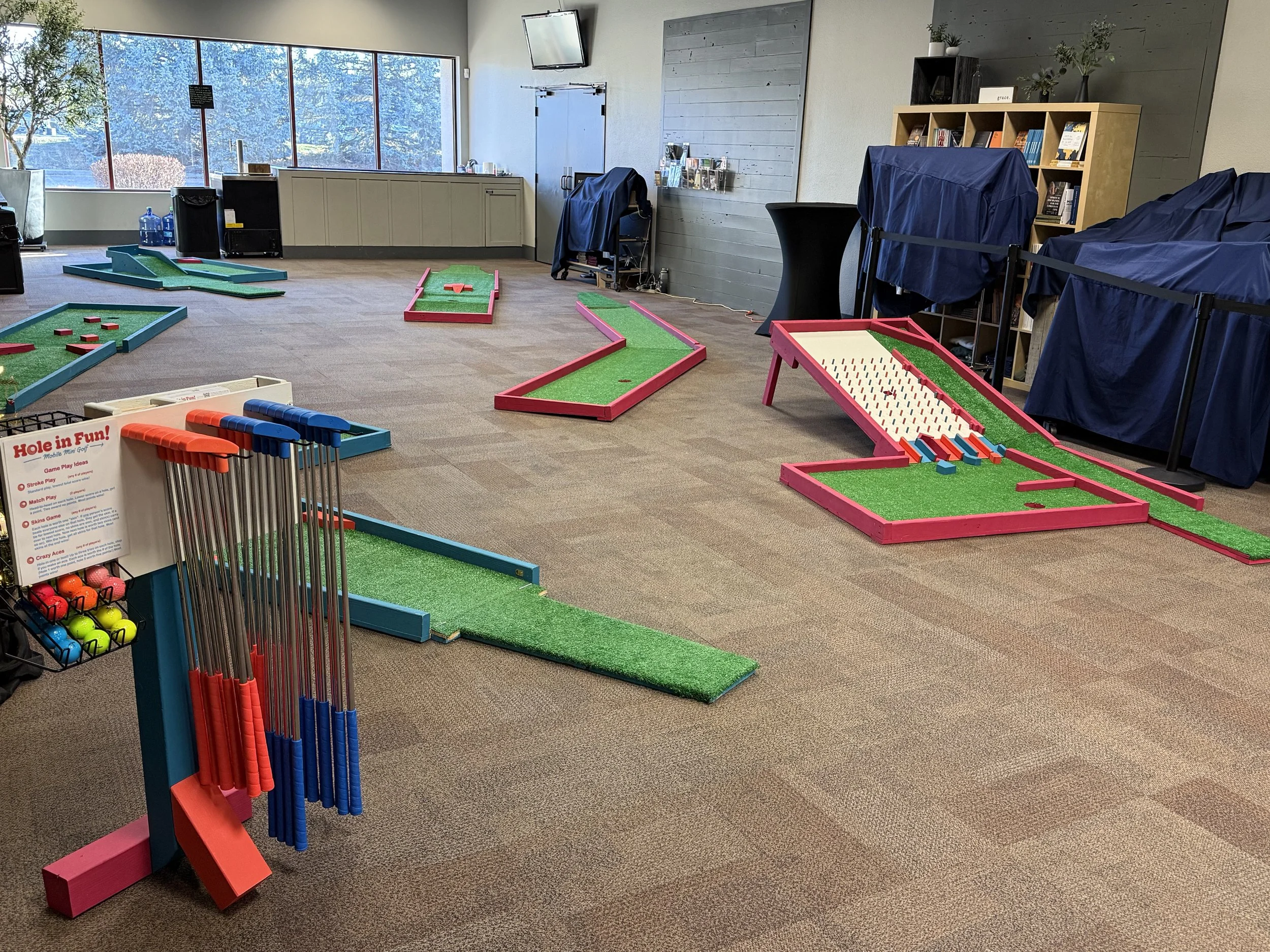 Indoor mini golf course with multiple holes and obstacles, golf clubs and balls at the front, large windows on the left side, and shelves with books and plants along the right wall.