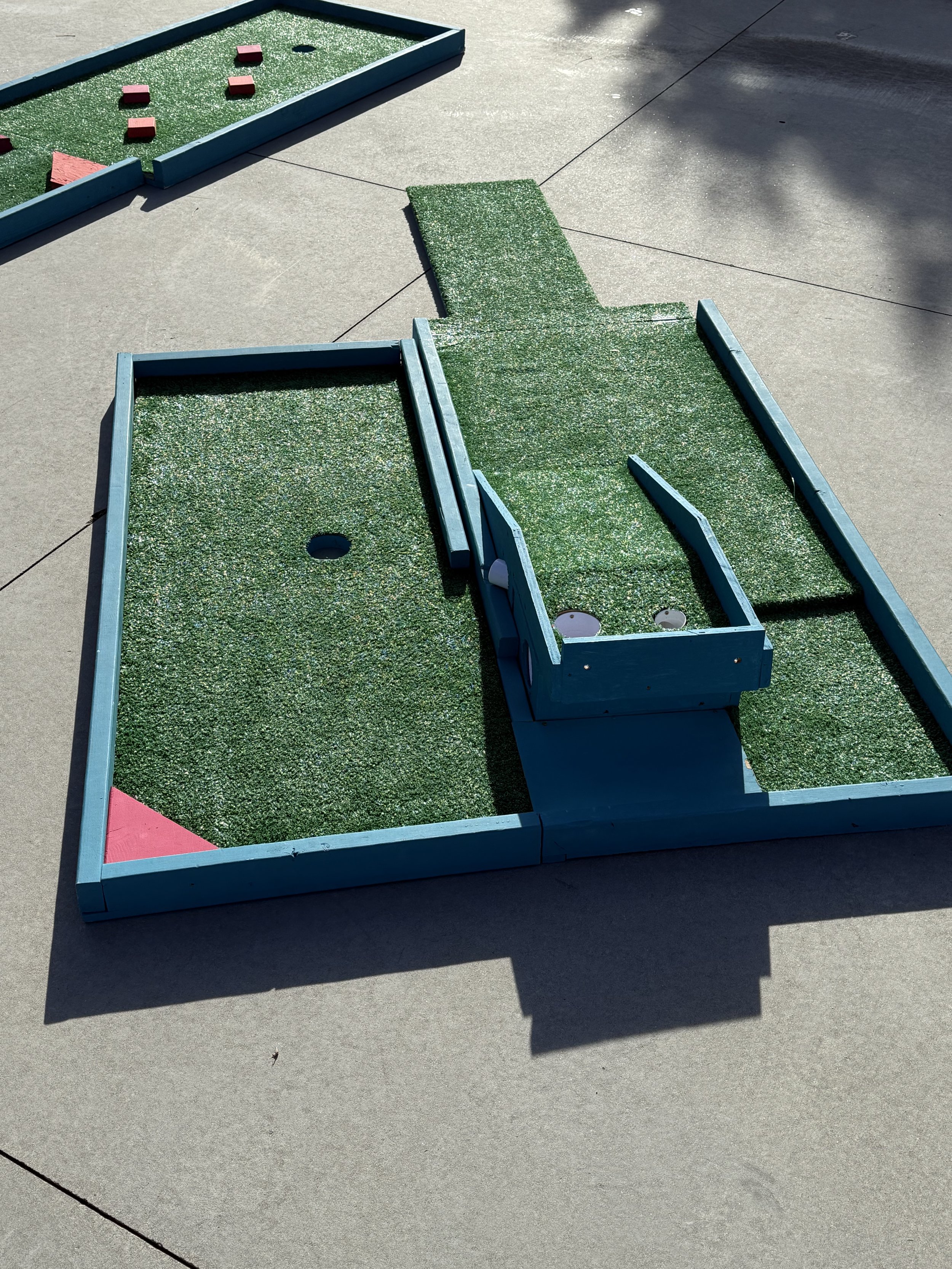 Disassembled outdoor mini golf putting green with blue wooden borders and artificial turf on a concrete surface.