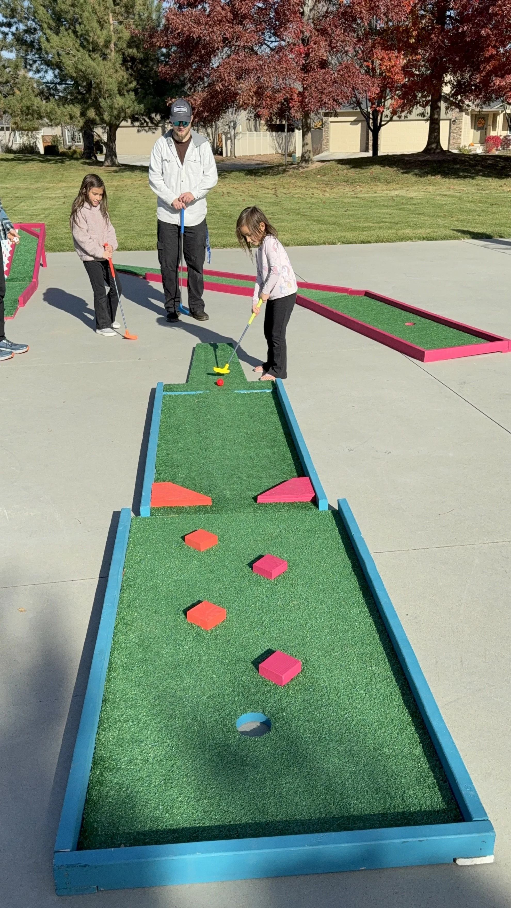 Children and an adult playing mini golf outdoors on a sunny day, with trees and houses in the background.