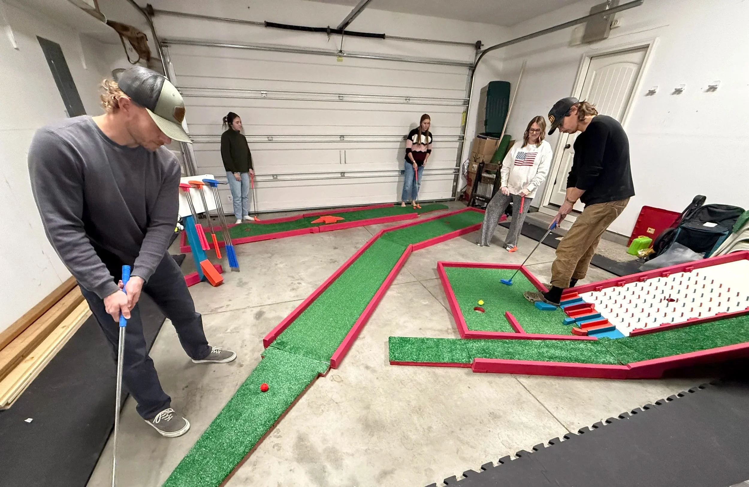 People playing miniature golf in a garage, with a small putting course featuring multiple holes, ramps, and obstacles.