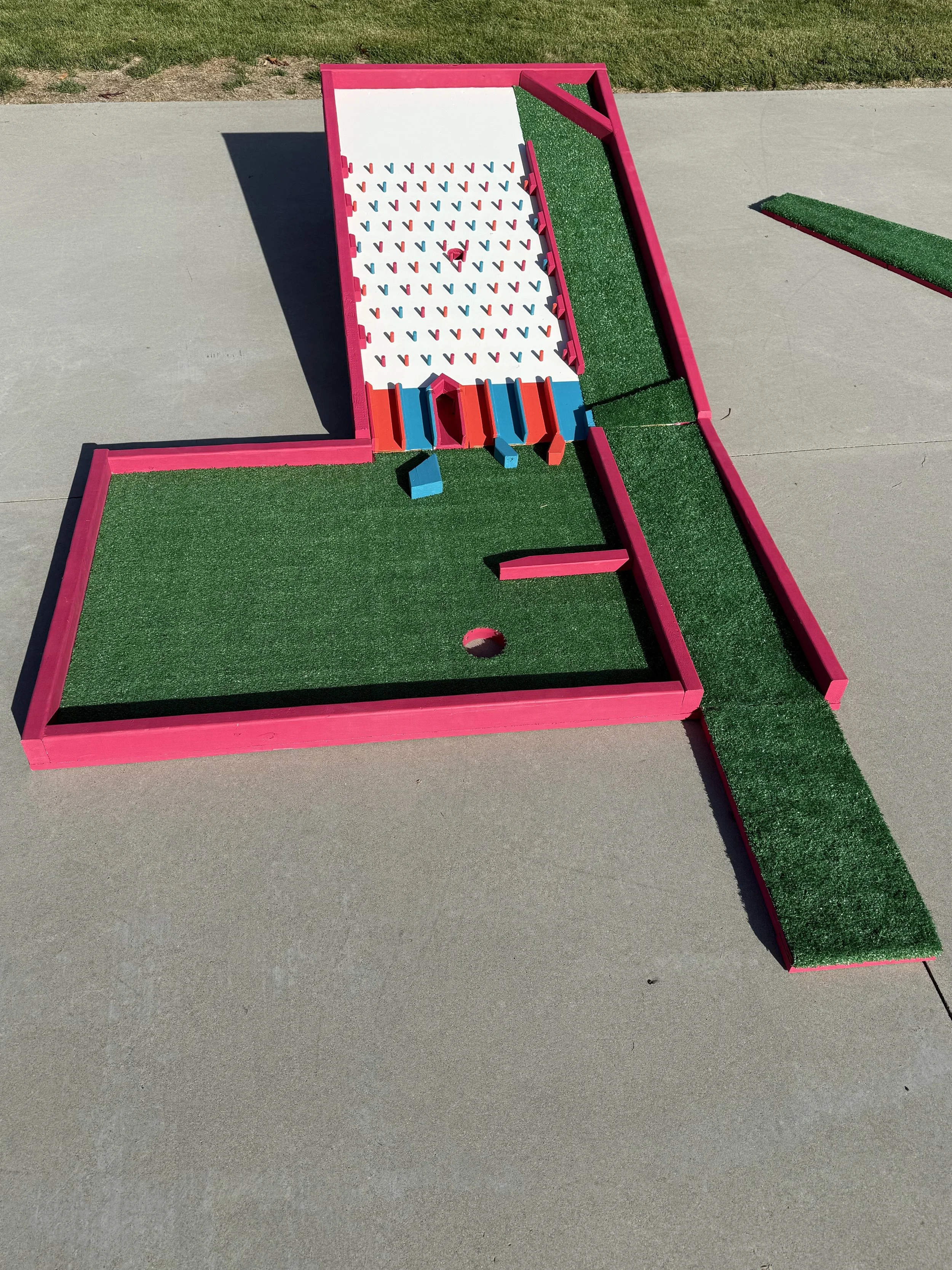 Miniature miniature golf course with pink borders, green artificial turf, and a small hole, set on concrete outdoors.