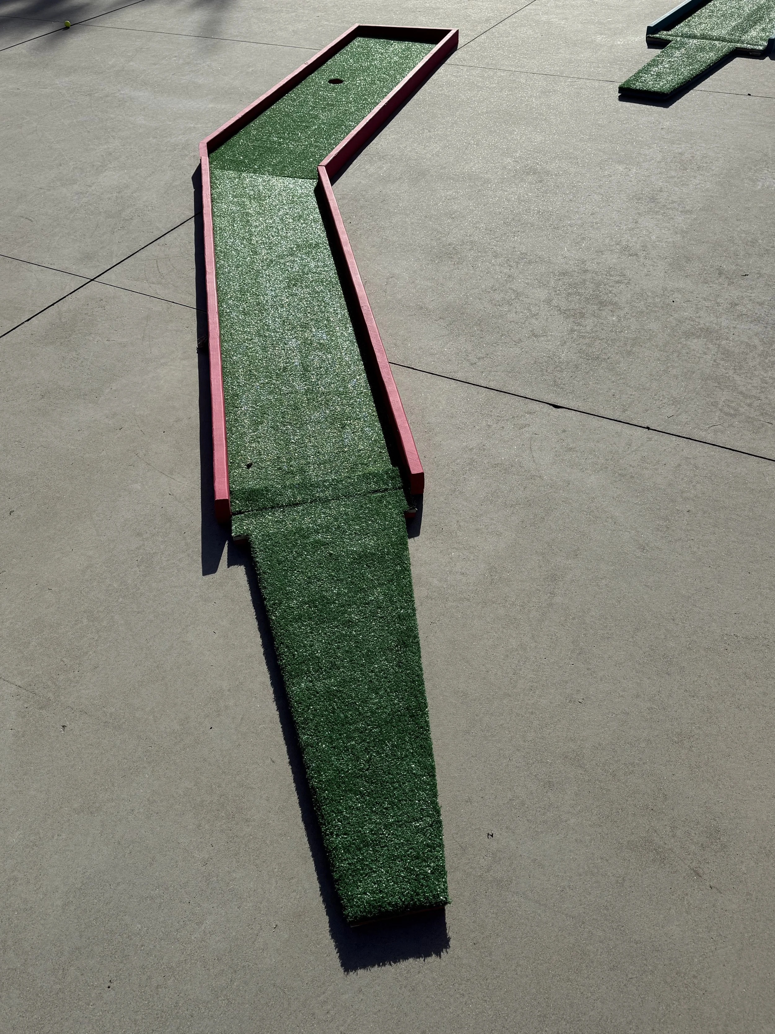 A mini golf putting green with a red border and green artificial turf, placed on a concrete surface.