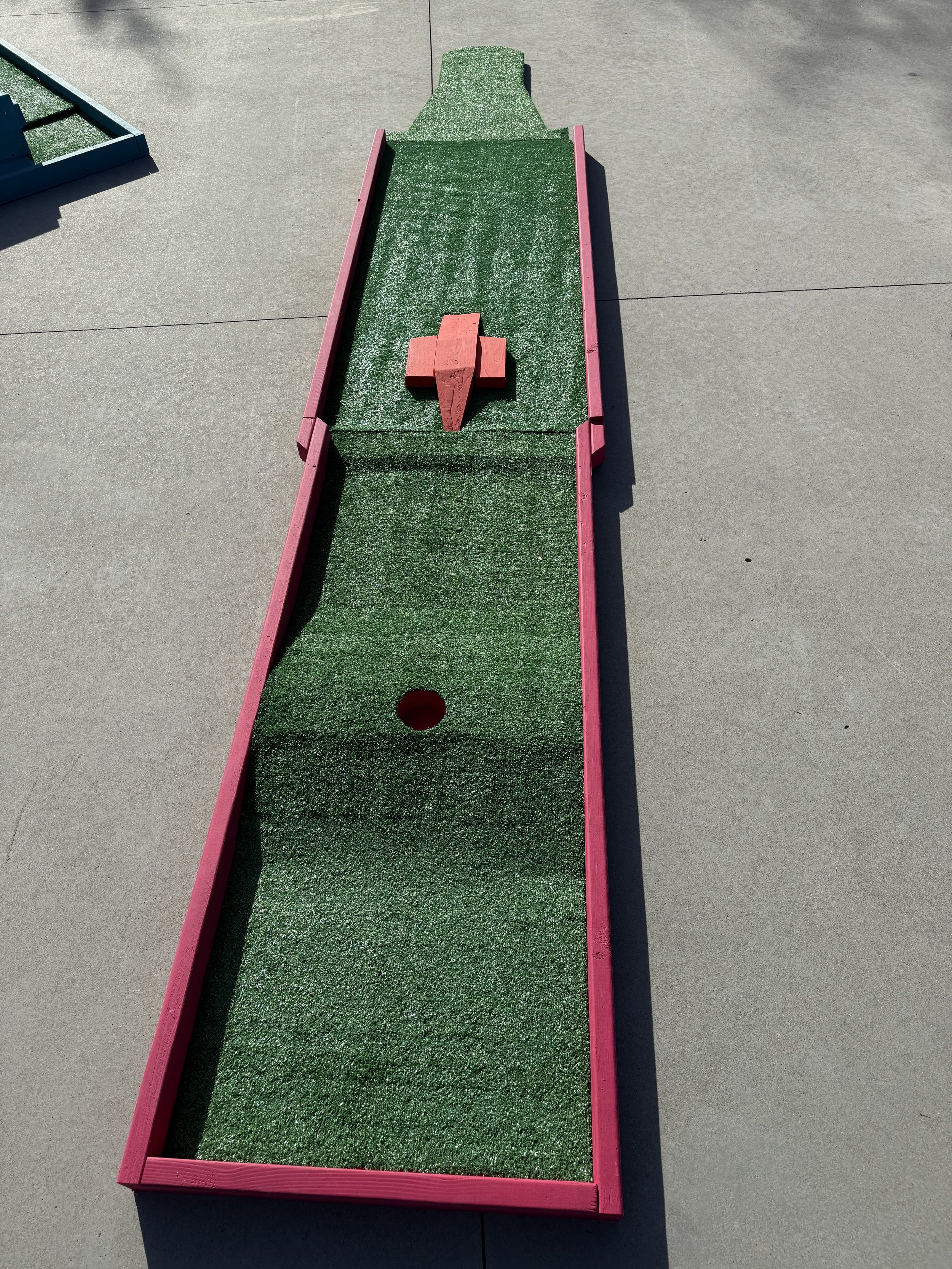 Miniature golf hole with green artificial turf, a red border, a red wooden bridge, and a red hole cup at the near end.