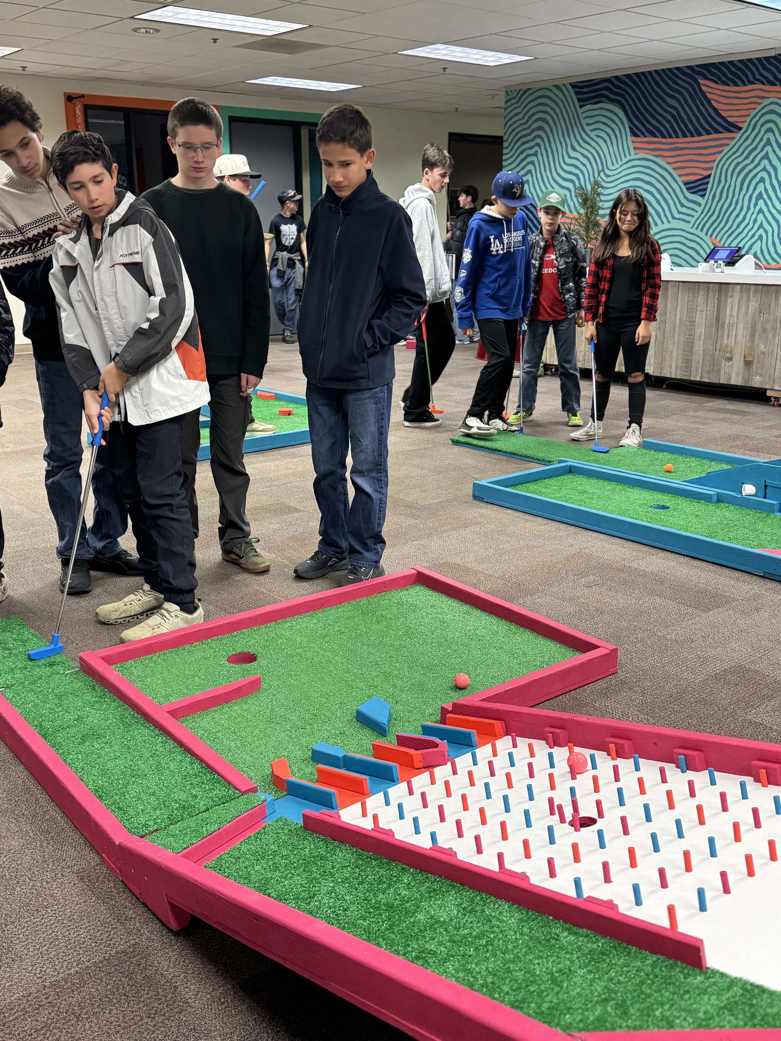 A group of children playing mini-golf indoors, with various colorful mini-golf holes and clubs, in a room with a patterned mural on the wall.