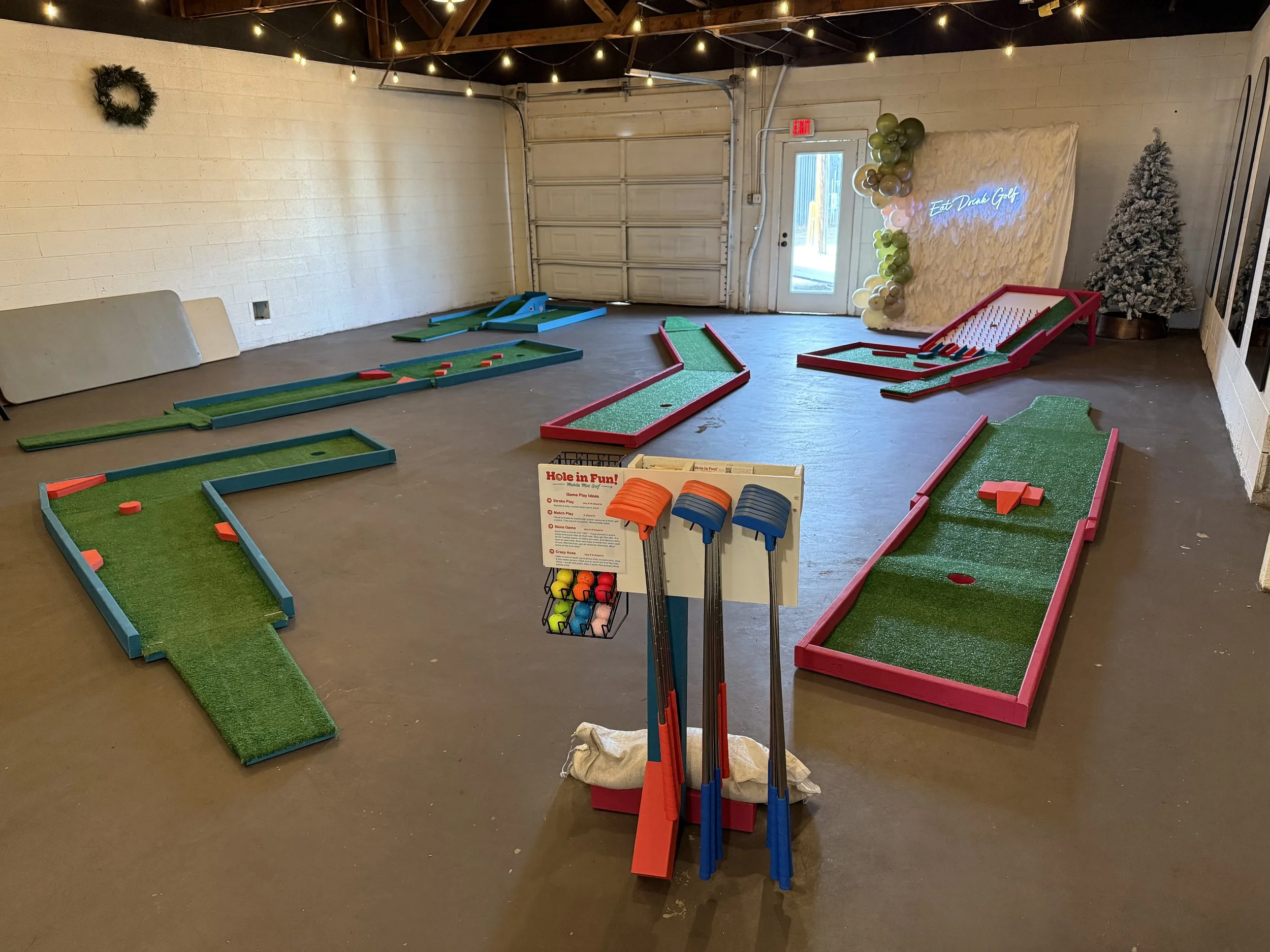 Indoor mini golf course with several small, colorful putt-putt holes arranged on the floor, a set of golf clubs and balls in the foreground, and holiday decorations including a light-up sign, a small white Christmas tree, and balloons in the backgrou