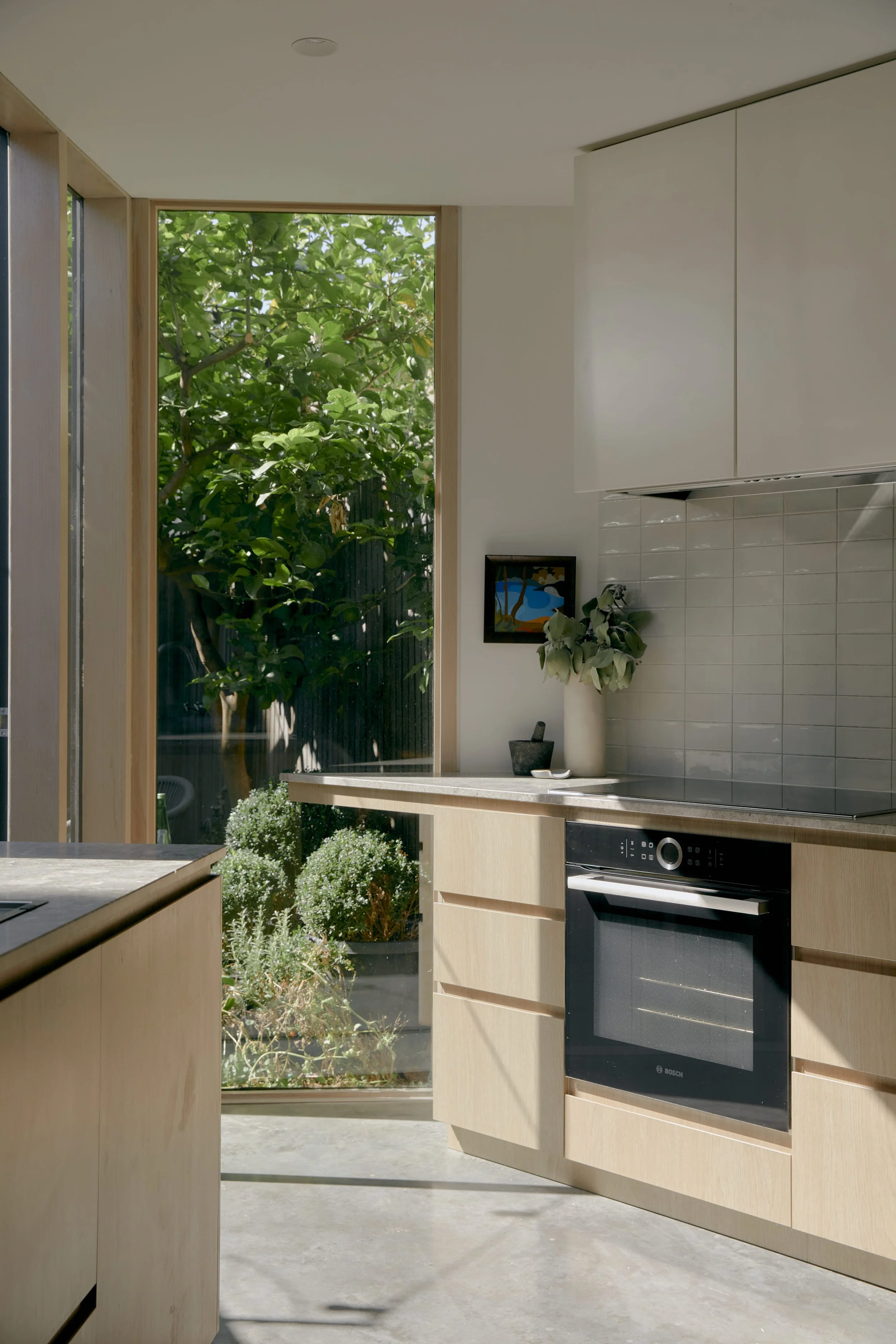 Kitchen with wooden cabinets, built-in oven, gray tiled backsplash, and large window showing green plants outside.