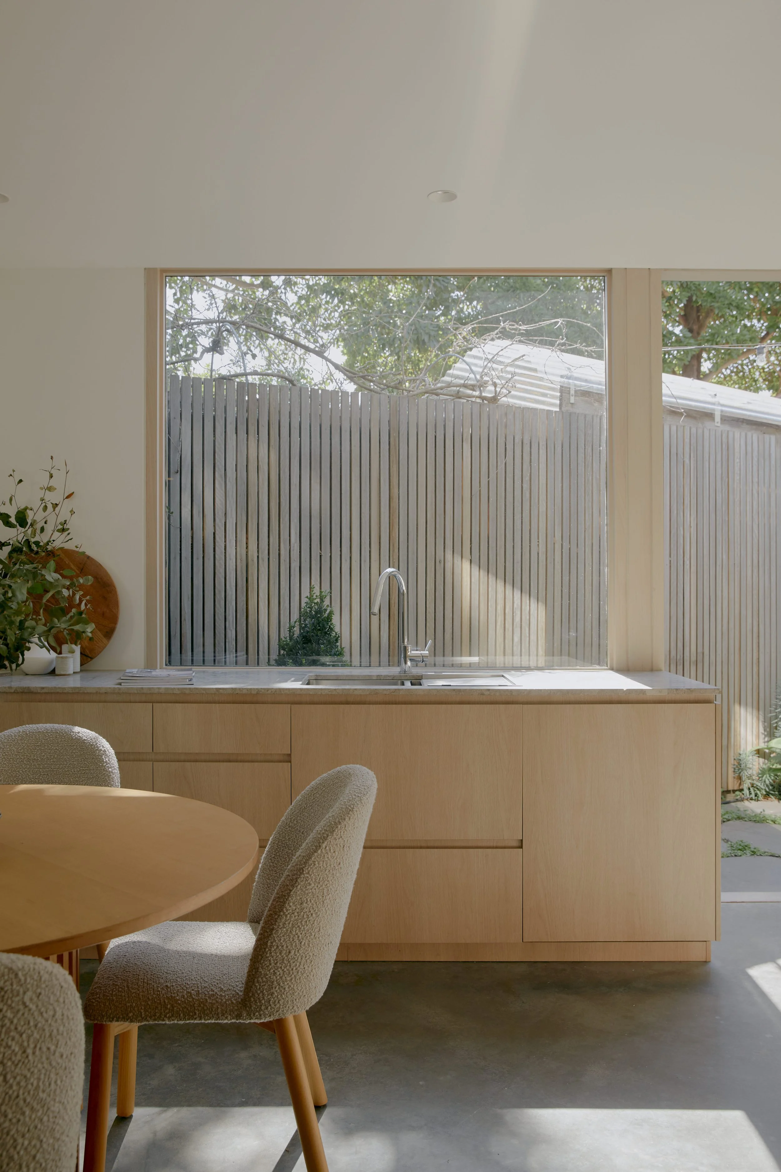 Bright kitchen with wooden cabinetry, a large window overlooking an outdoor wooden fence, a round wooden dining table with upholstered chairs, and sunlight streaming inside.