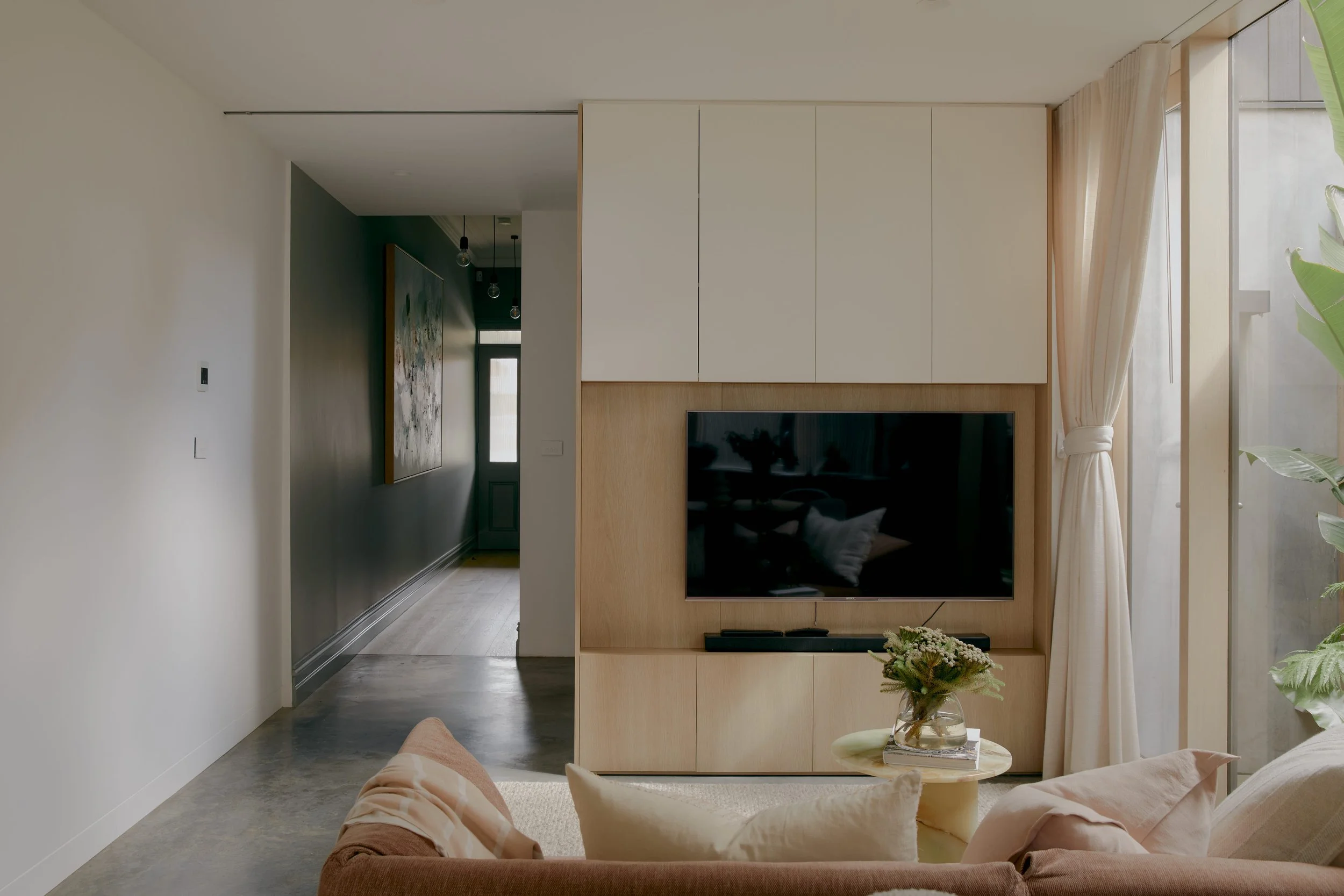 Living room with a mounted flat-screen TV on a light wood panel, beige and blush-colored sofa, a small round side table with a glass vase of flowers, and large windows with cream curtains letting in natural light.