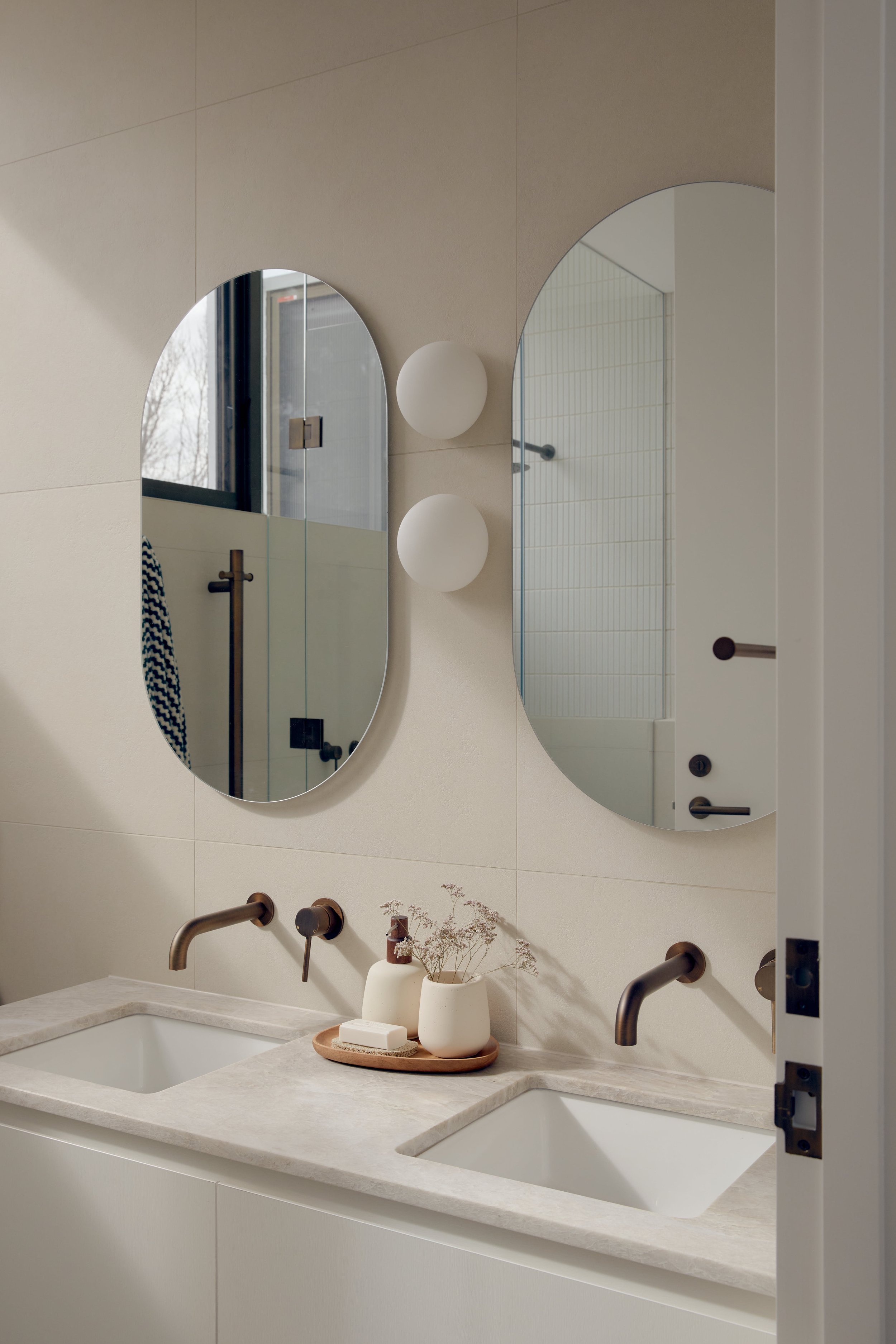 Modern bathroom with double sink, beige countertop, oval mirrors, white wall sconce lights, and minimal decor.