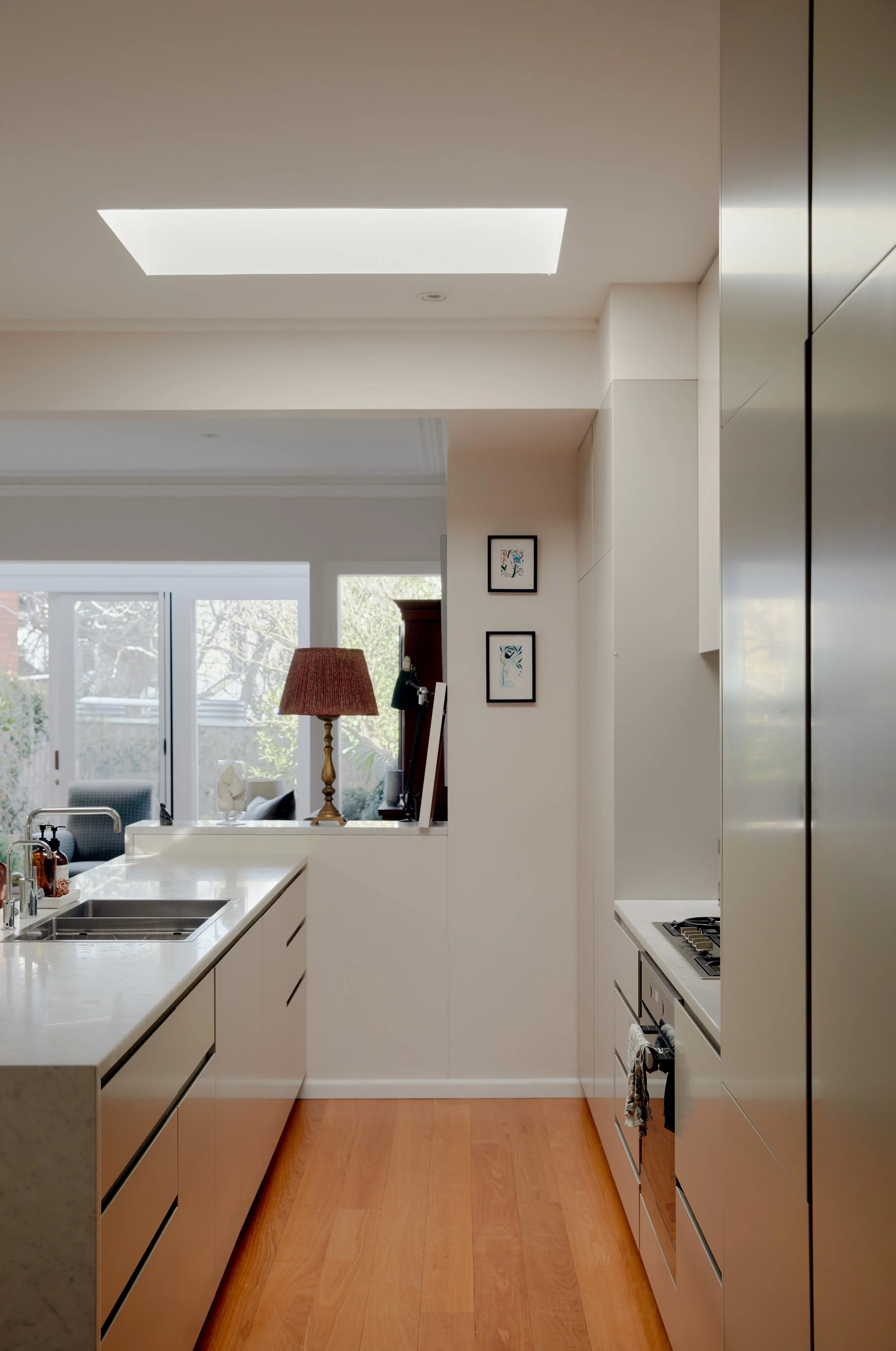 Modern kitchen with white cabinetry, stainless steel appliances, a wooden floor, and a view into a living room with windows and a sliding glass door, decorated with artwork and a lamp.