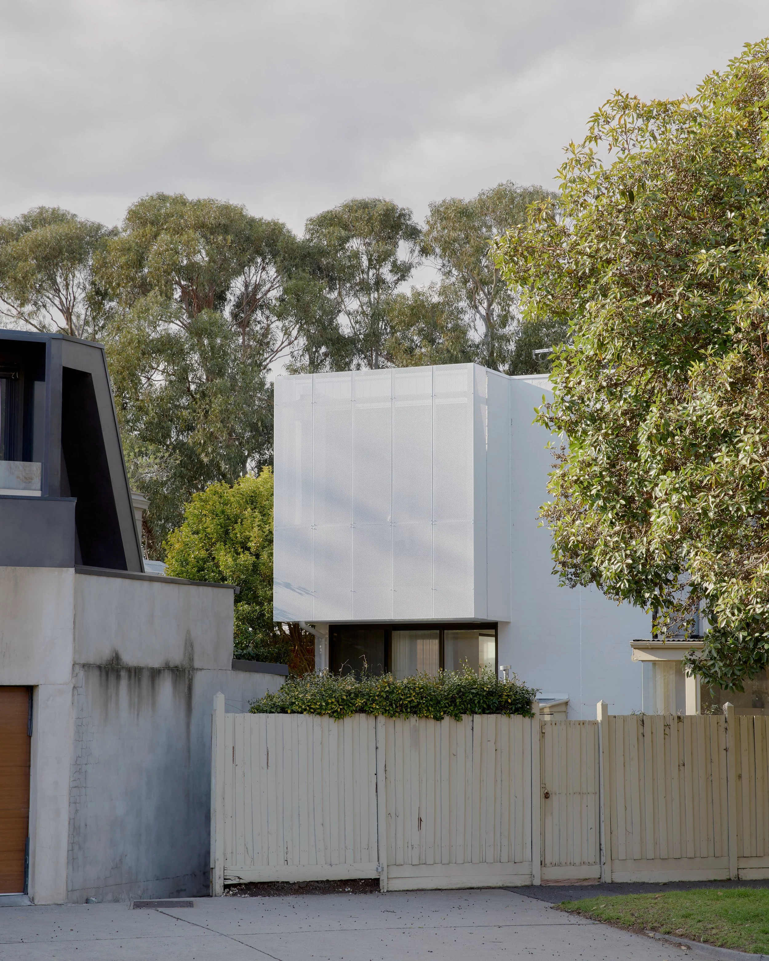 Modern white house with large window, surrounded by trees and a wooden fence.