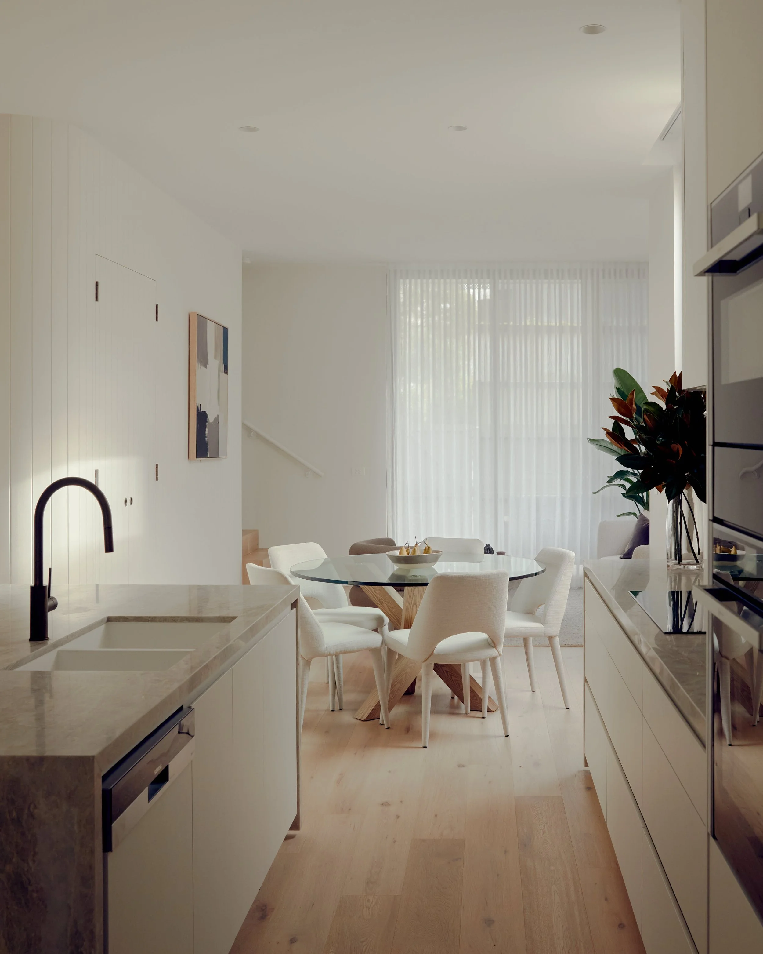 Modern, bright kitchen and dining area with a round table and white chairs, large window with sheer curtains, and light wood floors.