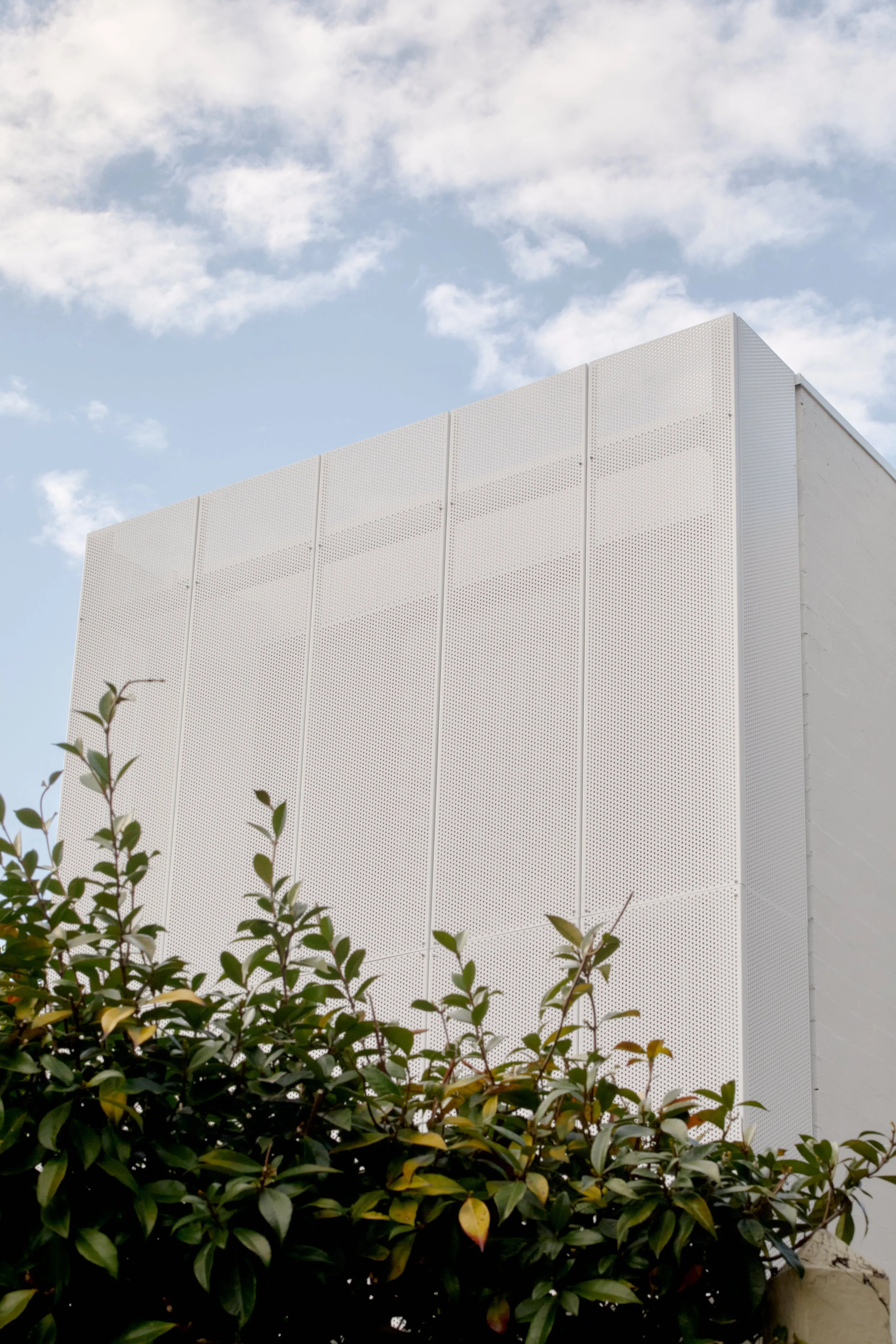 Close-up of a modern white building with a textured surface, against a blue sky with some clouds, and green bushes in the foreground.