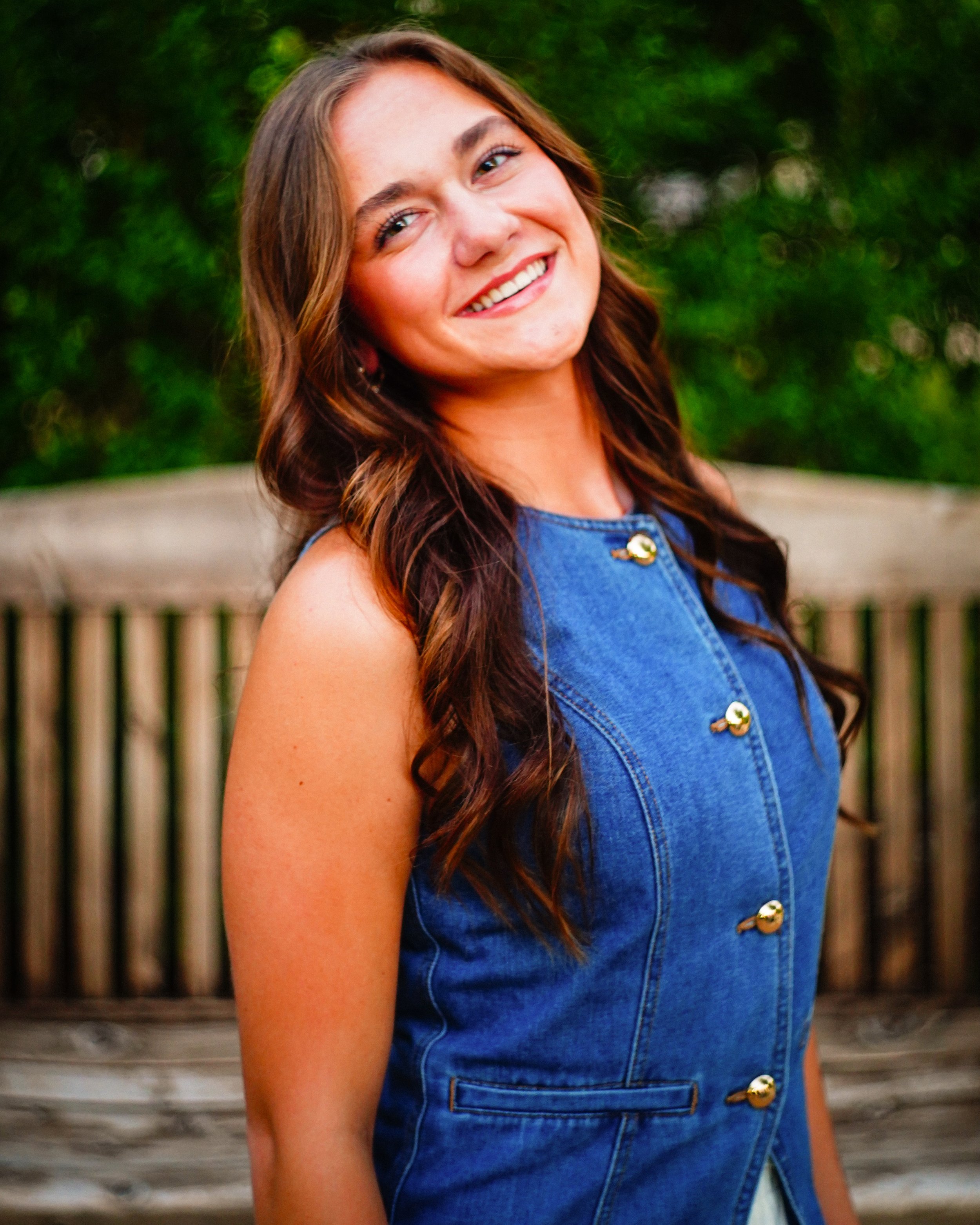 Close-up of a smiling woman with long, wavy brown hair wearing a sleeveless denim top, standing outdoors with a wooden fence and green foliage in the background.