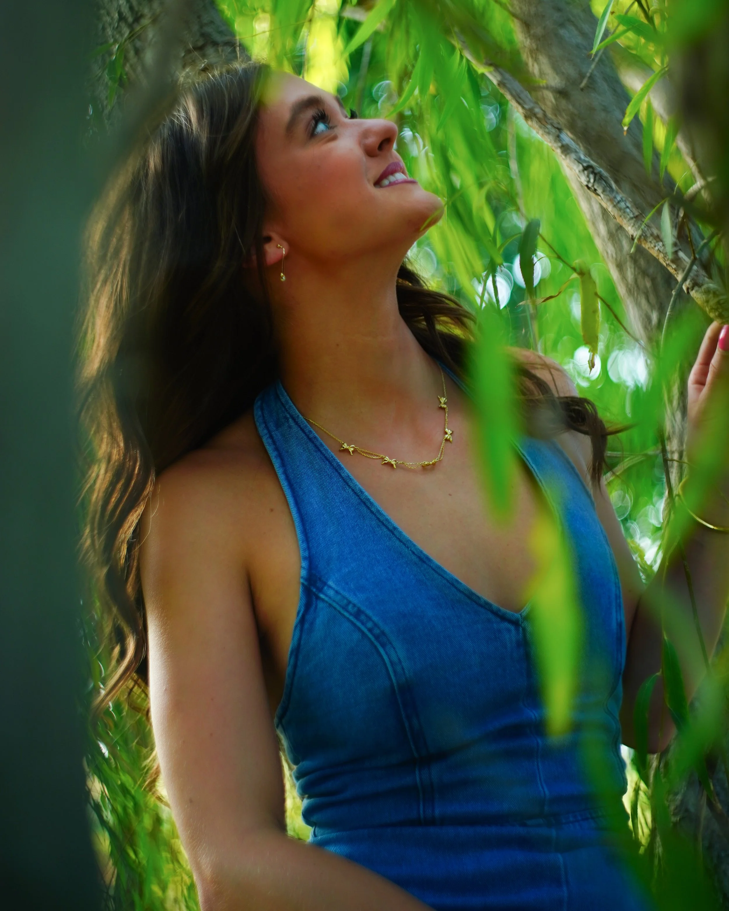 A woman with long brown hair and earrings looking up and smiling among green leaves and branches.