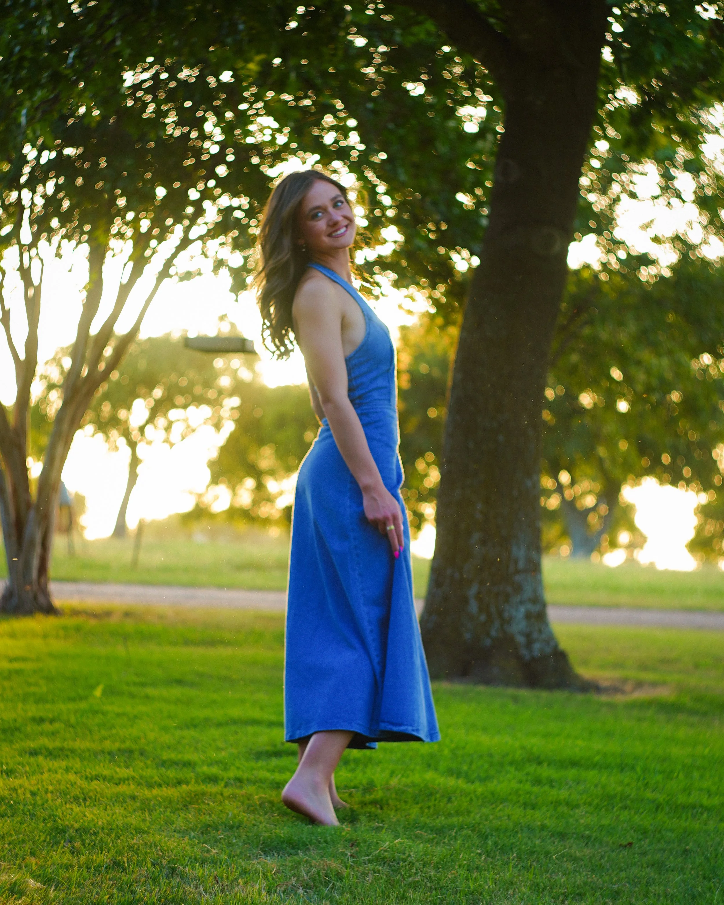 A woman in a blue dress standing outdoors in a grassy area, near trees, with sunlight in the background, smiling at the camera.
