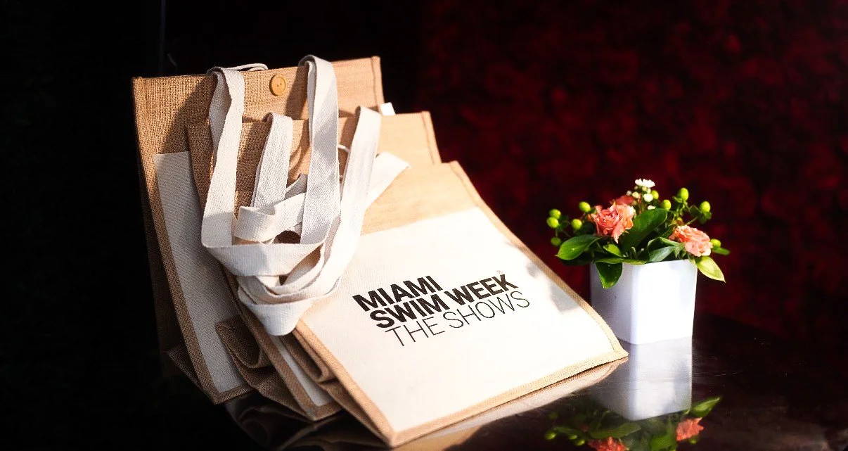 A tote bag with text "Miami Swim Week The Shows" next to a white pot of pink and peach flowers on a dark surface.