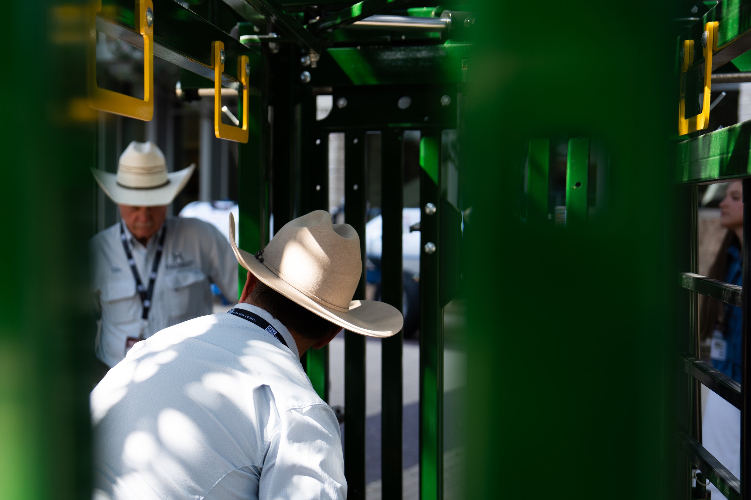 People interacting near a green structure, with one person wearing a wide-brimmed hat and others wearing hats or helmets, at an outdoor event or exhibition.