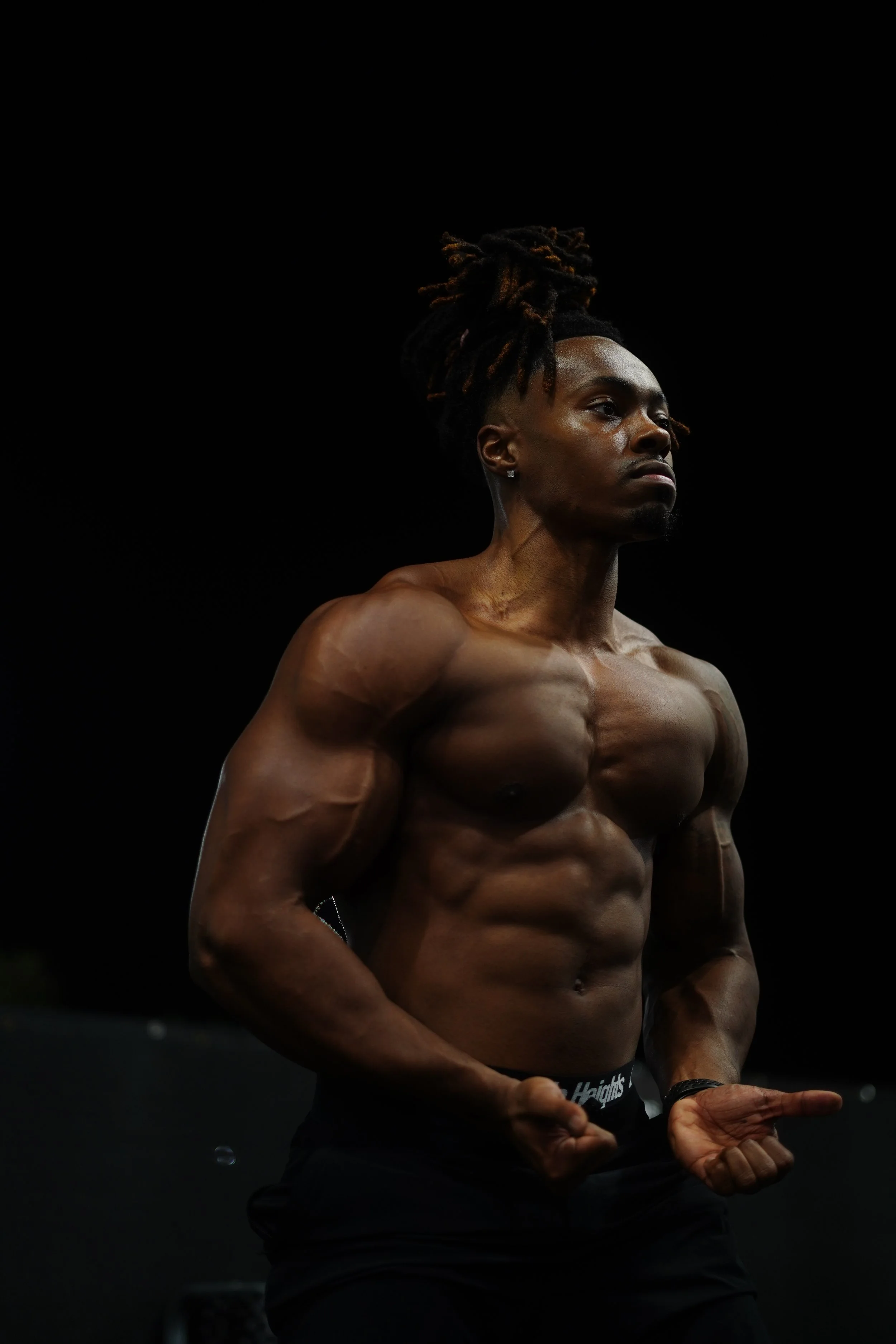 A shirtless male athlete with a muscular build and dreadlocks, standing against a dark background, flexing his arms and showing defined abs.