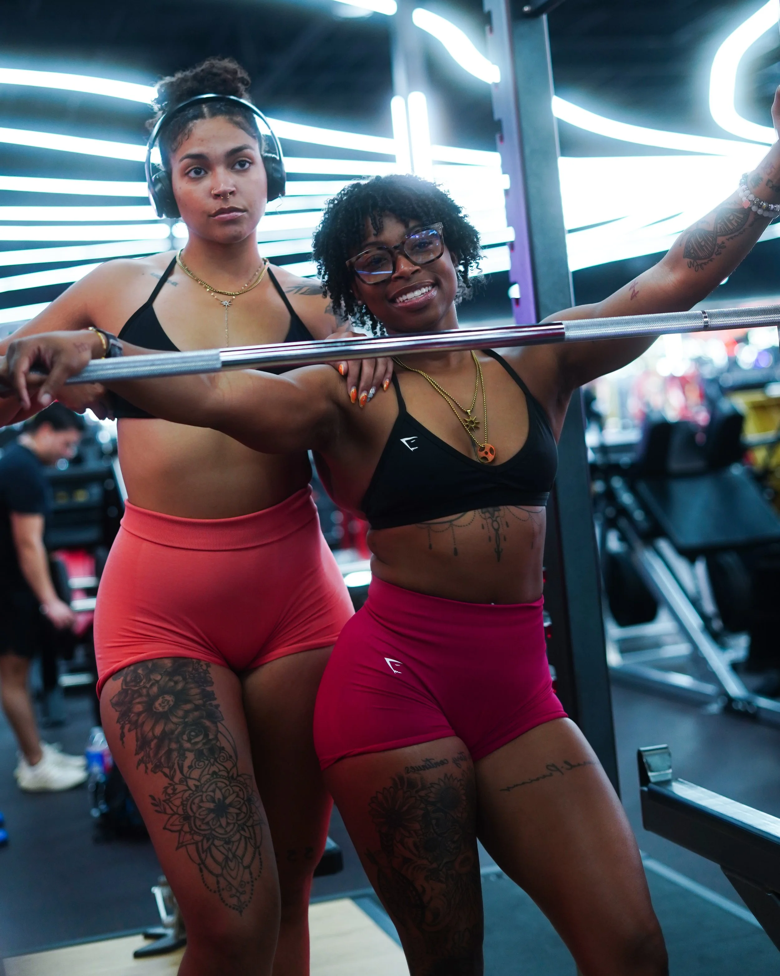 Two women with tattoos and tattoos, wearing black sports bras and red shorts, working out in a gym with neon lights and exercise equipment in the background.