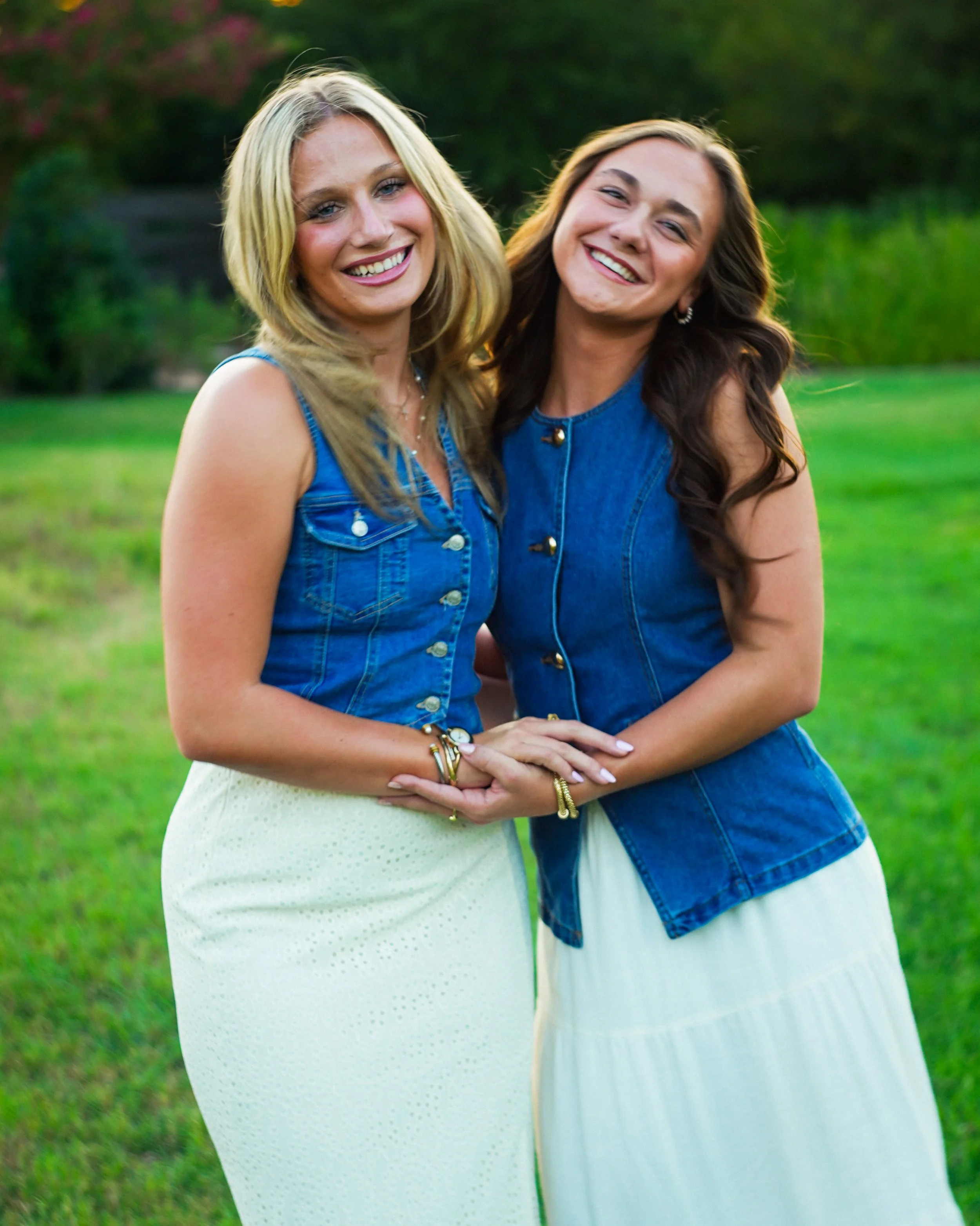 Two women smiling and standing close together outdoors with greenery in the background, wearing blue denim vests over white dresses.