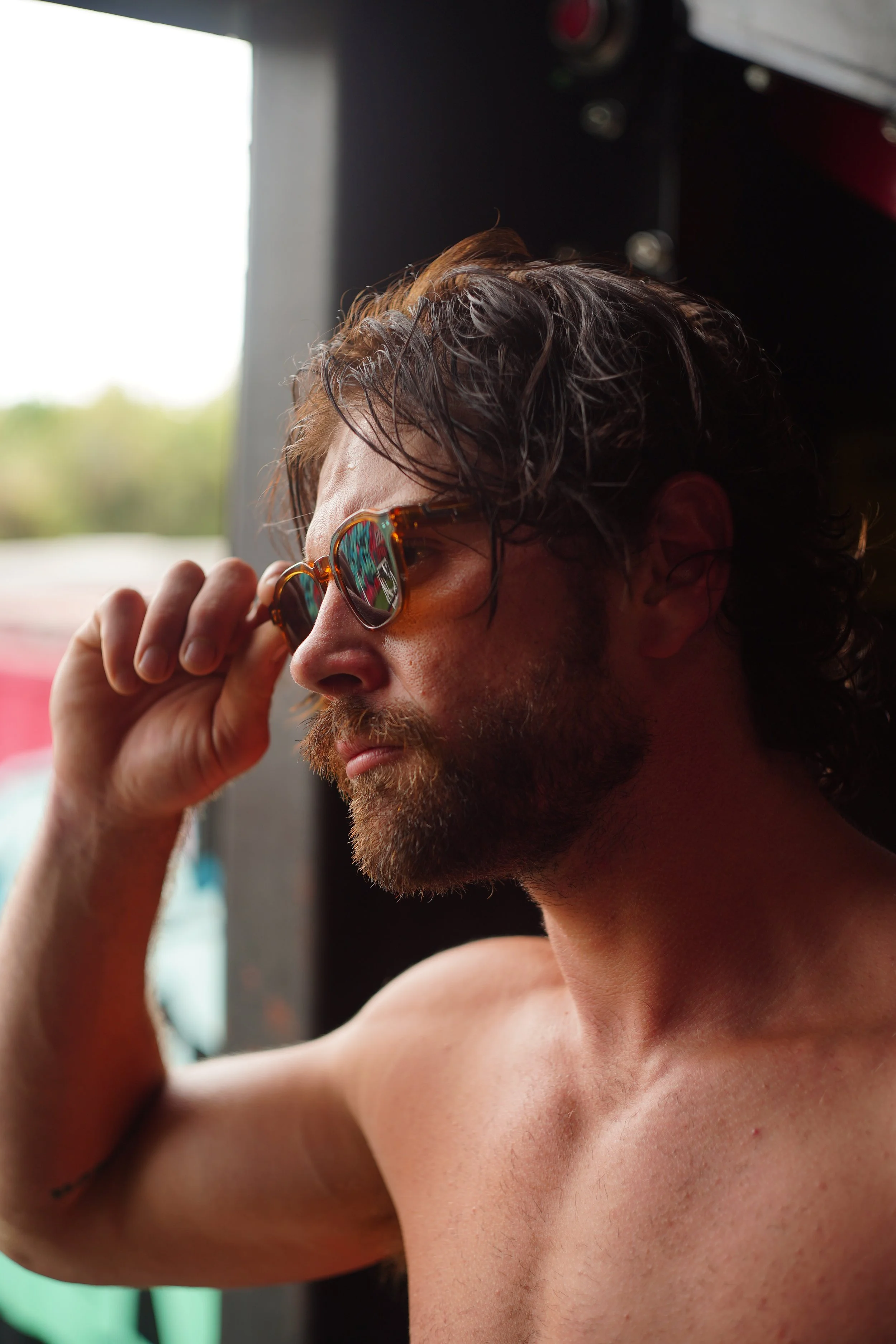A shirtless man with glasses adjusting them with his hand, sitting indoors near a window.