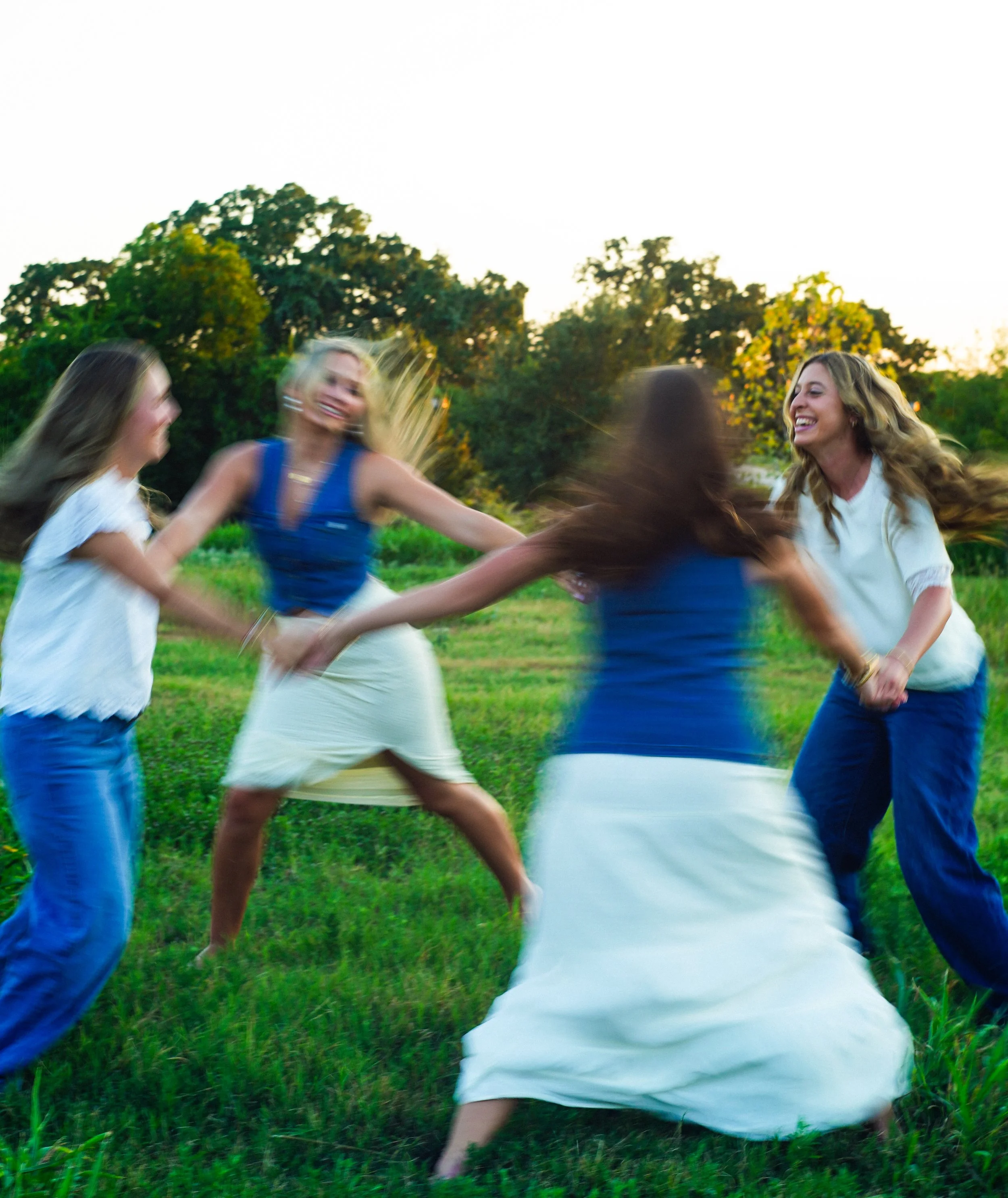 A group of women holding hands, forming a circle, outdoors on grass with trees in the background, during sunset or late afternoon. They are smiling and appear to be enjoying themselves.
