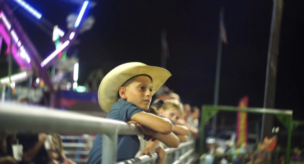 A young boy wearing a cowboy hat leans on a railing at a nighttime carnival with brightly lit rides and other children in the background.