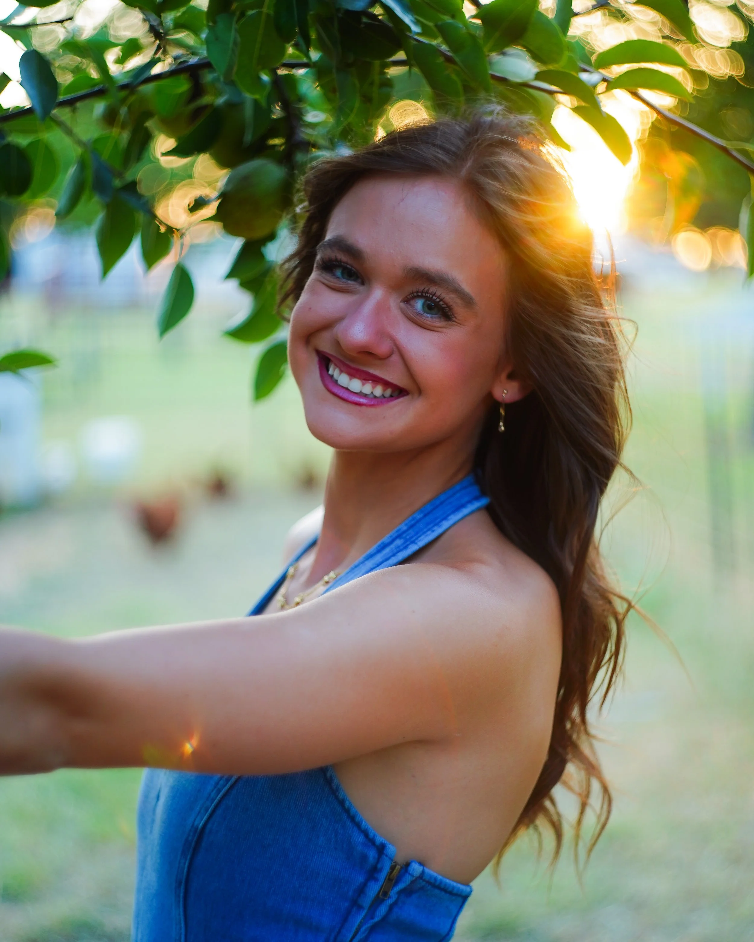 A smiling young woman with curly brown hair and blue eyes, wearing a blue sleeveless top and earrings, standing outdoors in a park or garden during sunset, with lush green trees and sunlight creating a warm glow.
