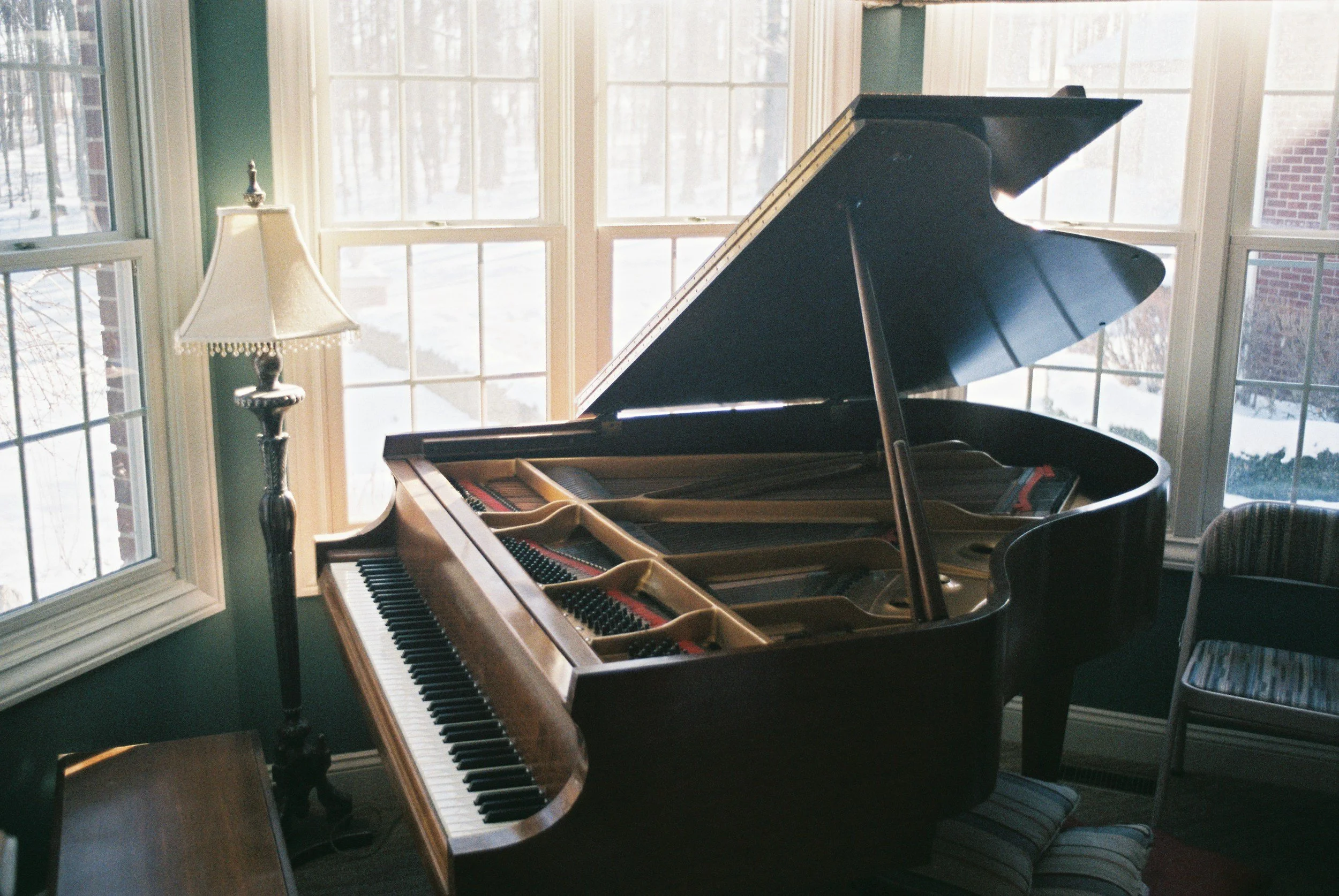A black grand piano with an open lid in a room with large windows showing a snowy outdoor scene. There is a standing lamp with a beige shade next to the piano and a chair with striped upholstery nearby.
