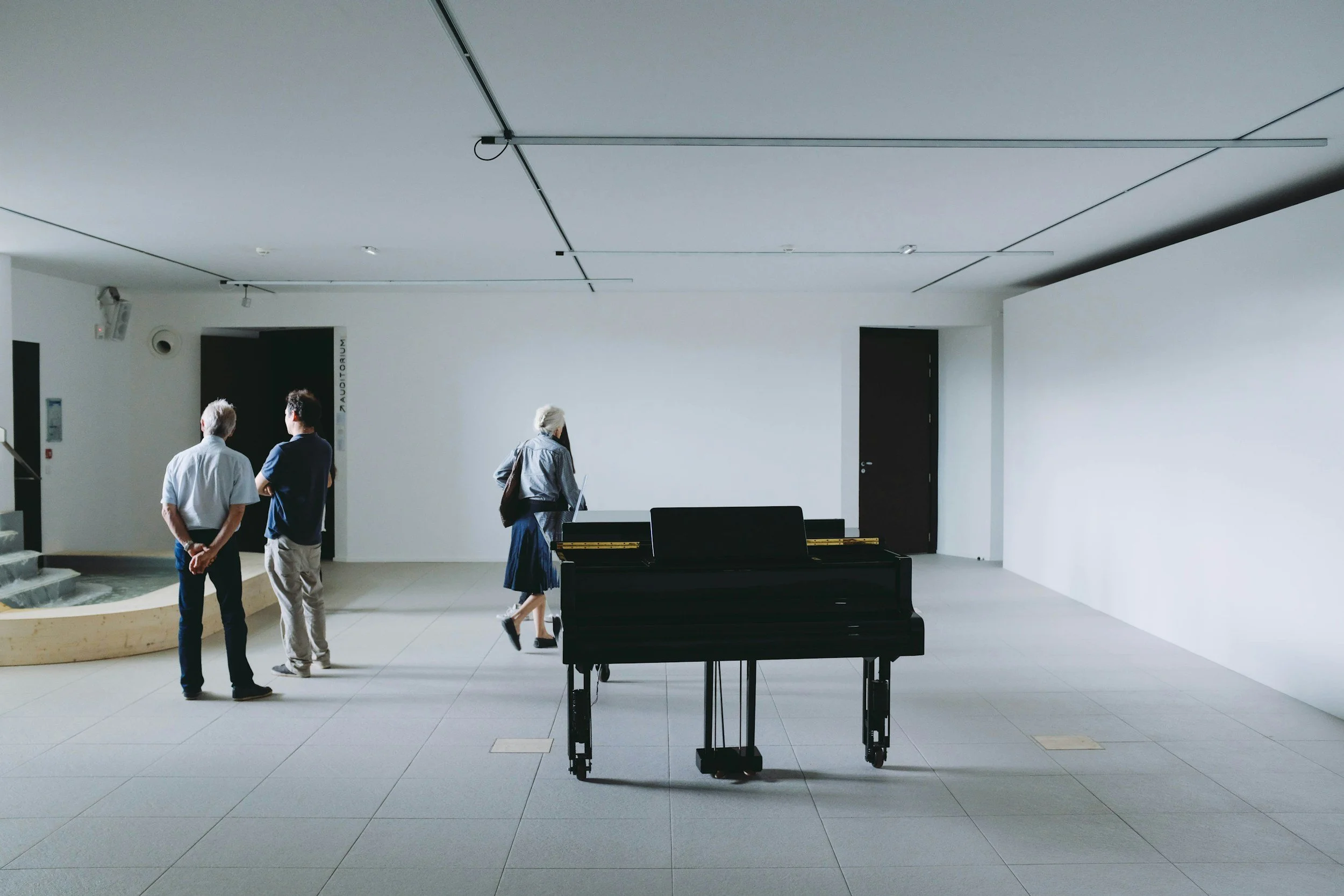 An empty black grand piano stands in the center of a white-walled room with three people walking and standing nearby, and a small staircase on the left.