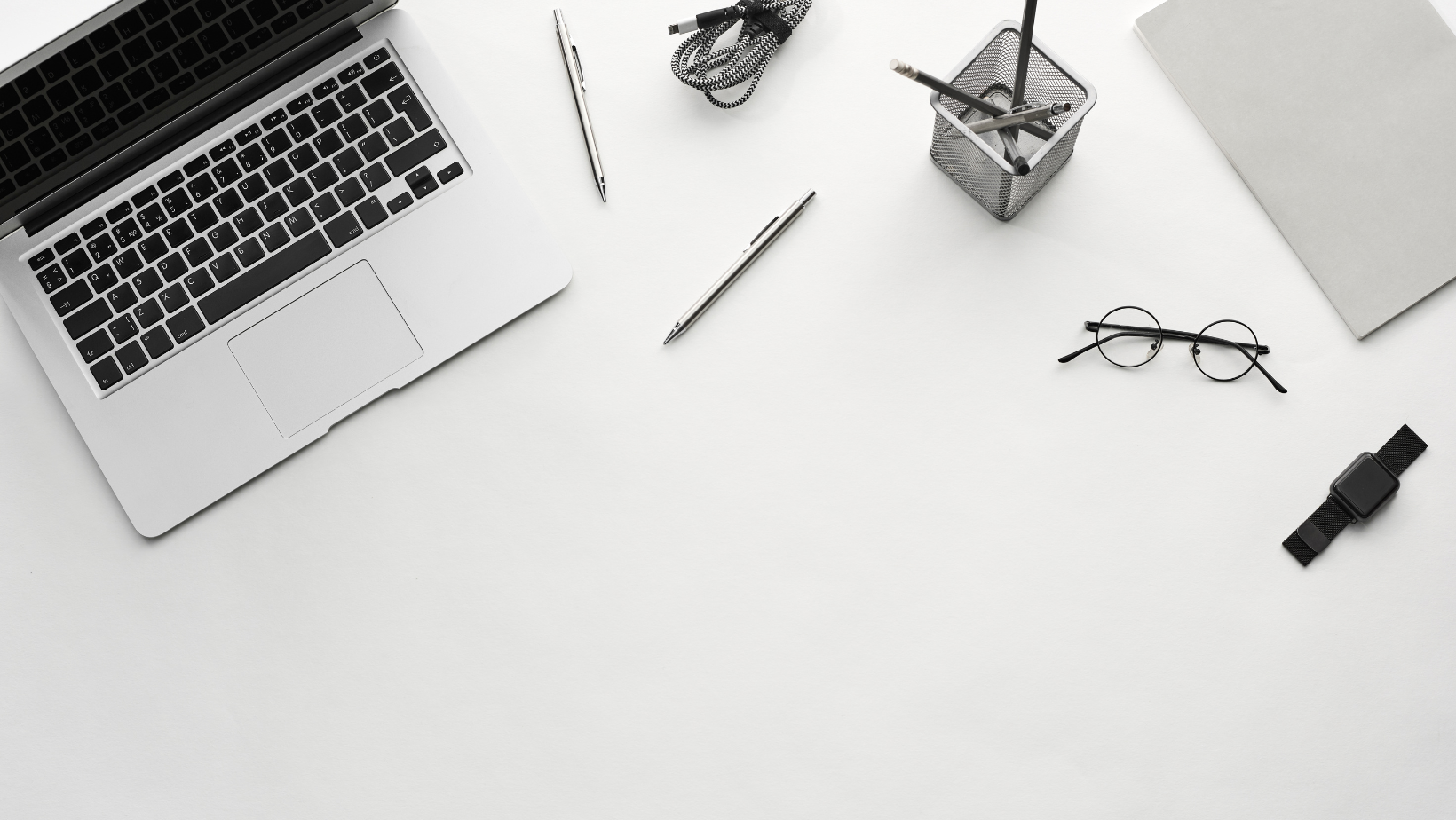White desk with a silver laptop, two pens, a pair of glasses, a black smartwatch, a notebook, a small mesh container with pens, and a gray cloth.