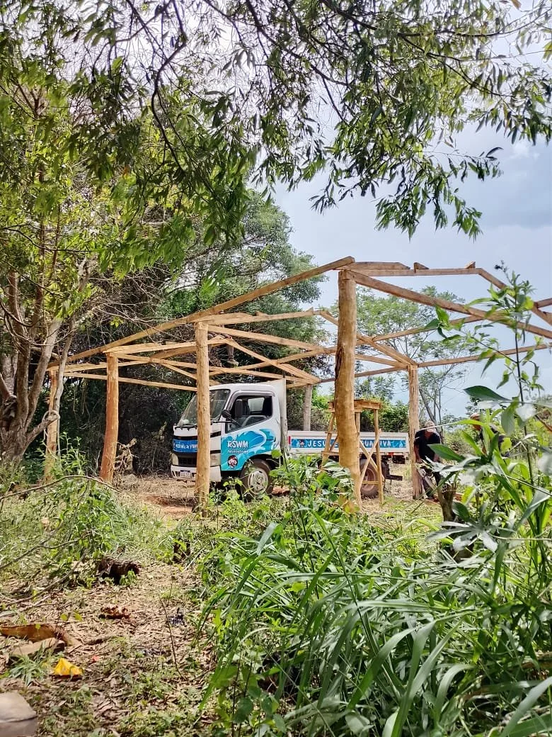 A small truck is parked in a natural, wooded area with trees and greenery. The truck has text and graphics on its side, and wooden poles and framework are under construction around it, likely for a shelter or structure.