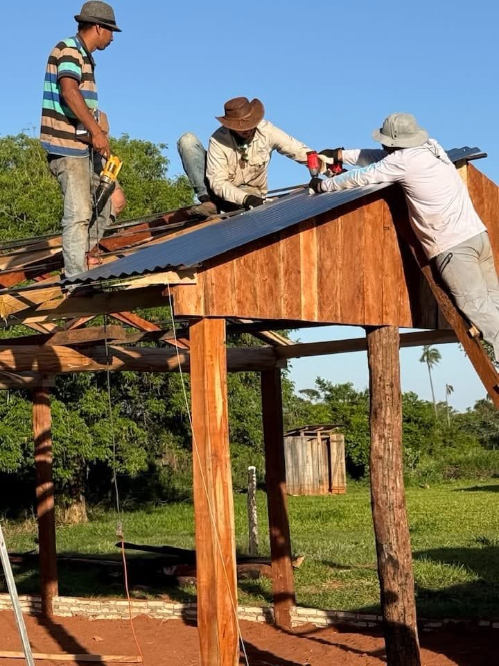 Three men working together on a wooden roof, with one standing, one kneeling on the roof, and another leaning against the roof. They are using tools and wearing hats for protection, under a clear blue sky.