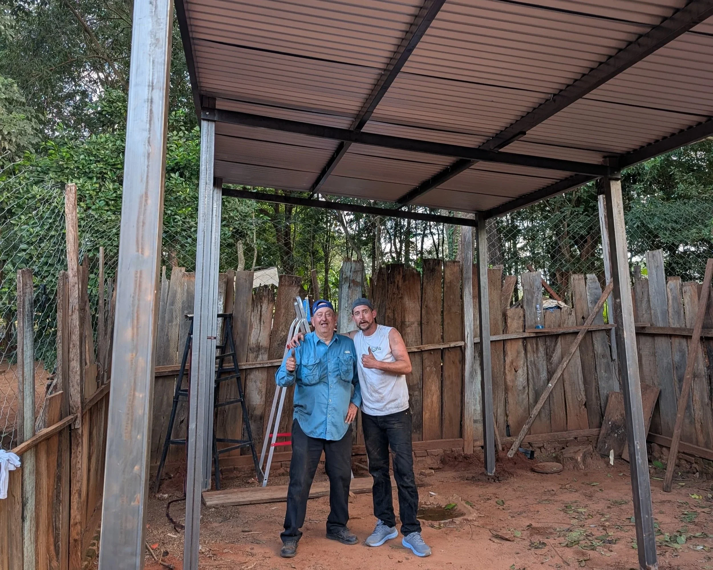 Two men standing inside a partially built outdoor structure with a metal roof, wooden walls, and a dirt ground, smiling and giving thumbs-up signs.