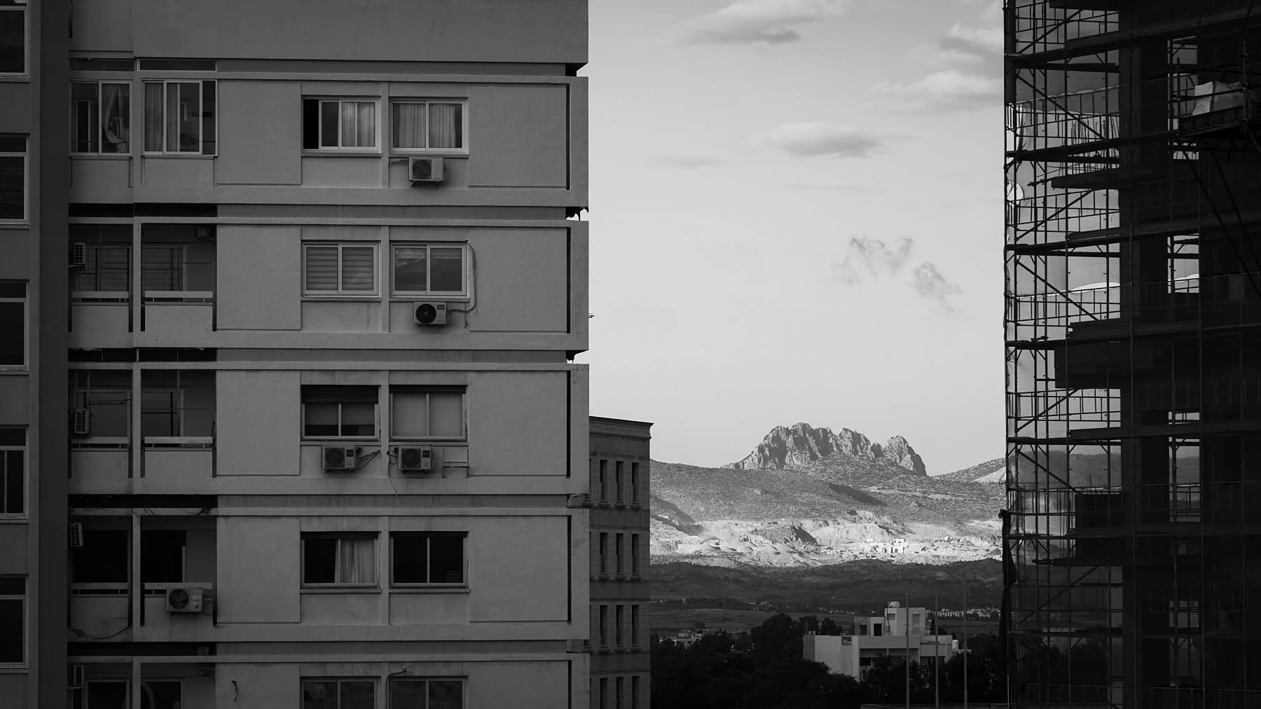Black and white photo showing two buildings, one with balconies and air conditioning units on the left, and the other under construction with scaffolding on the right. In the background, mountains and a partly cloudy sky are visible.