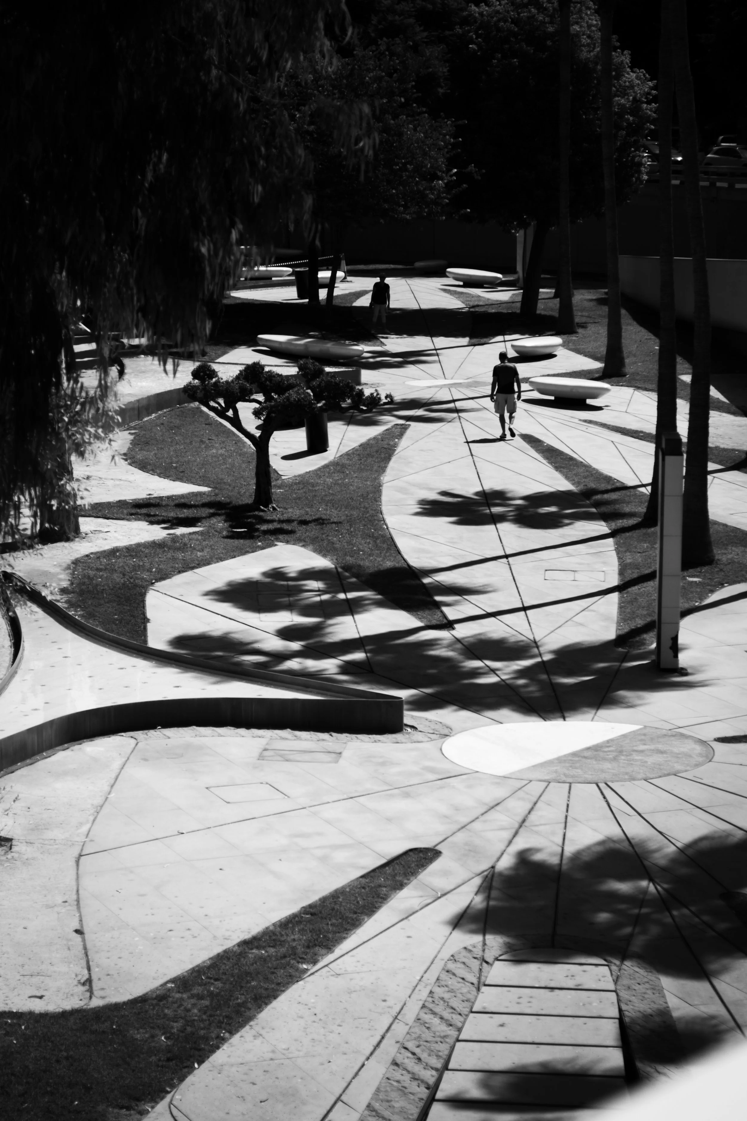 Black and white photo of a park with a winding path, small trees, and benches. Two people are walking along the path, casting shadows. Tall trees line the area, and there are some parked cars visible at the top right.