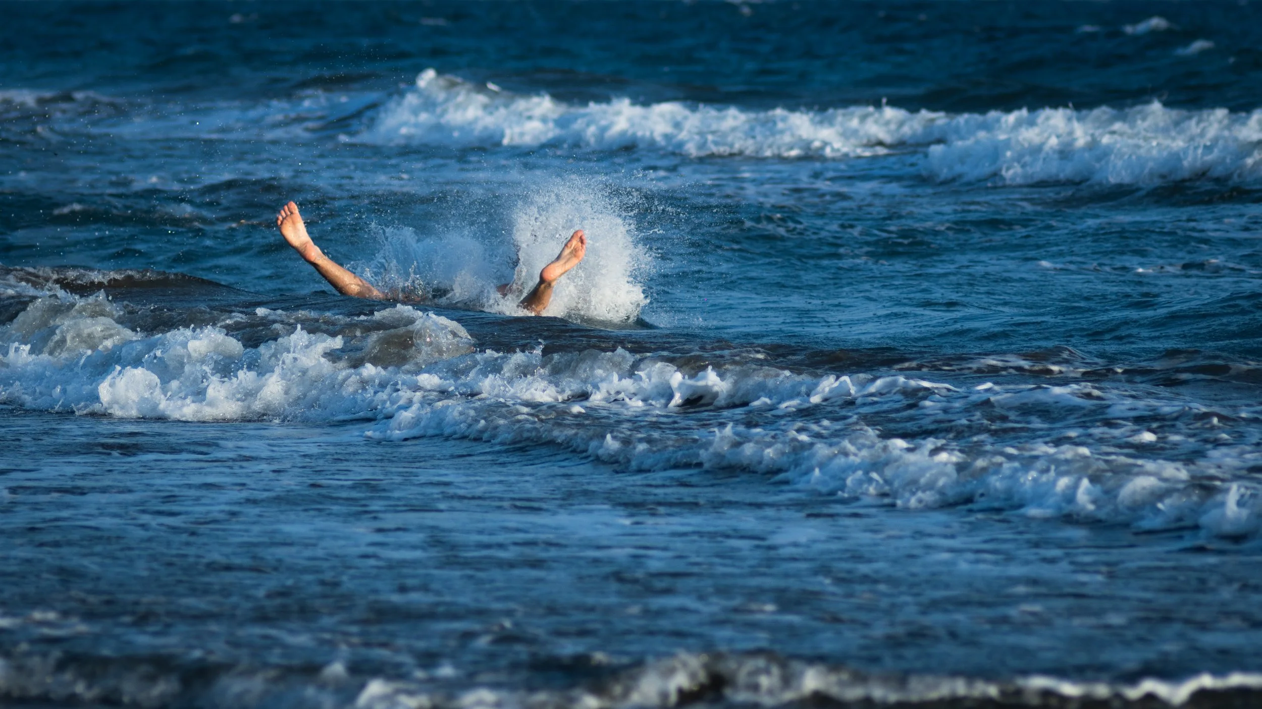Person falling into the ocean with arms and legs visible above the water, surrounded by waves and foam.