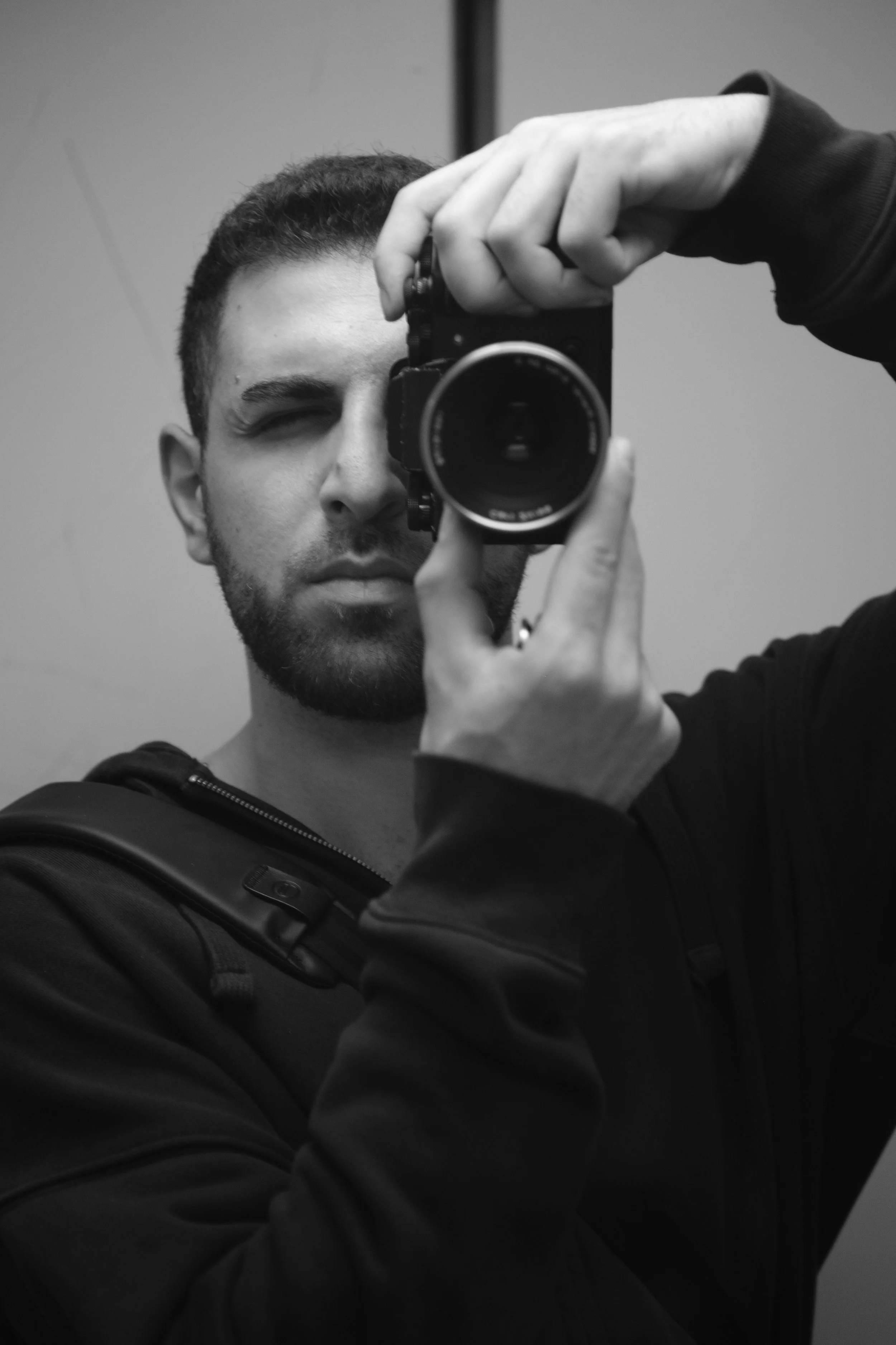 A man taking a selfie with a camera, in black and white, wearing a dark hoodie, with short hair and a beard.