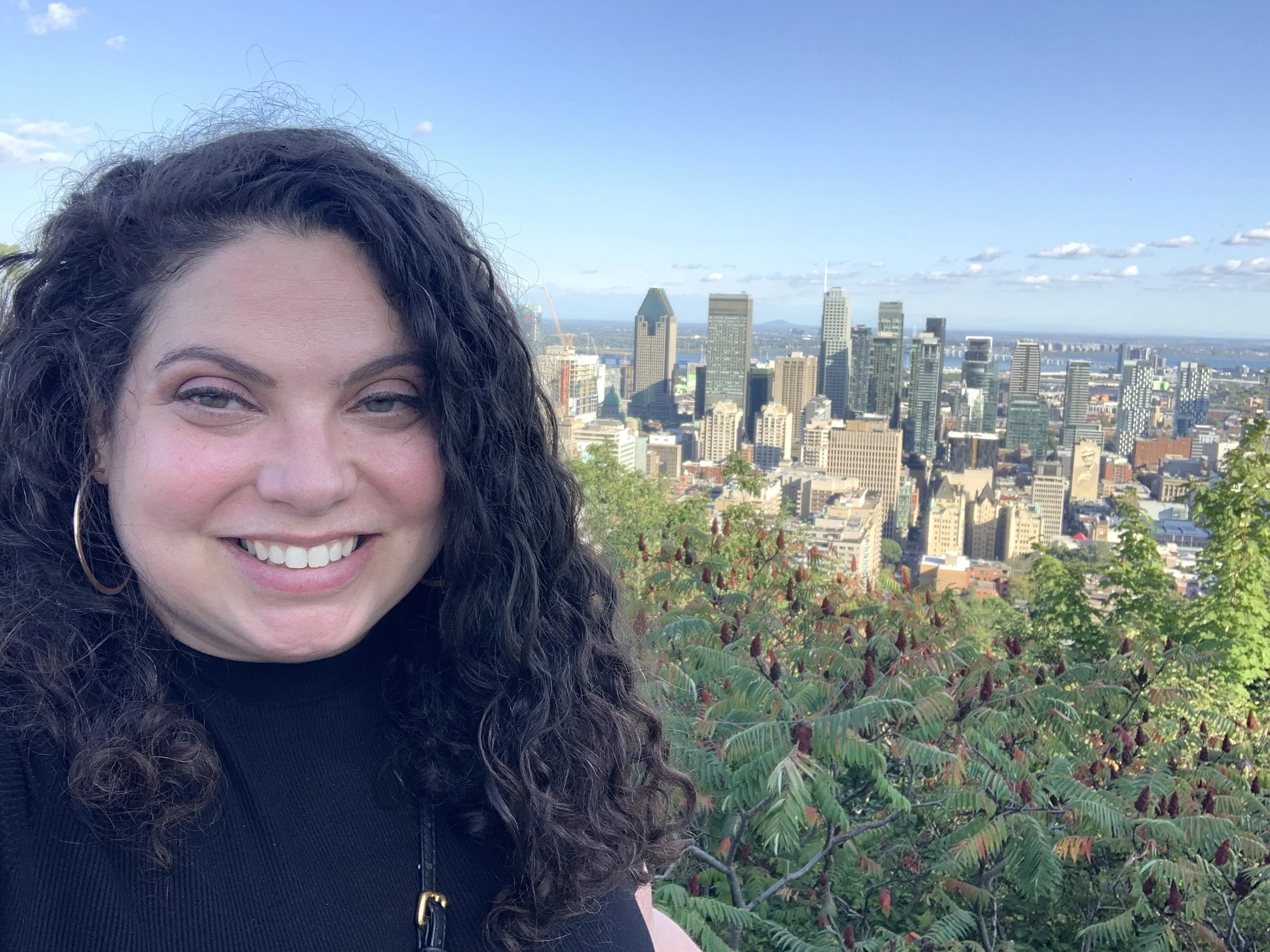 A woman with curly dark hair smiling, with a city skyline in the background, including tall skyscrapers and greenery in the foreground.