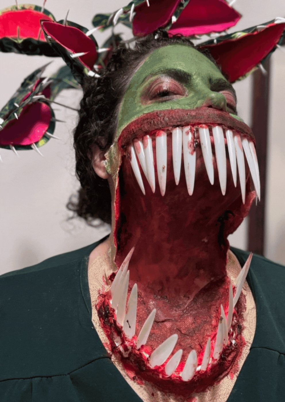 Person in a costume with a large, monster-like mouth with sharp teeth, spikes, and red accents, and green face paint, wearing an elaborate headdress with red and pink elements.
