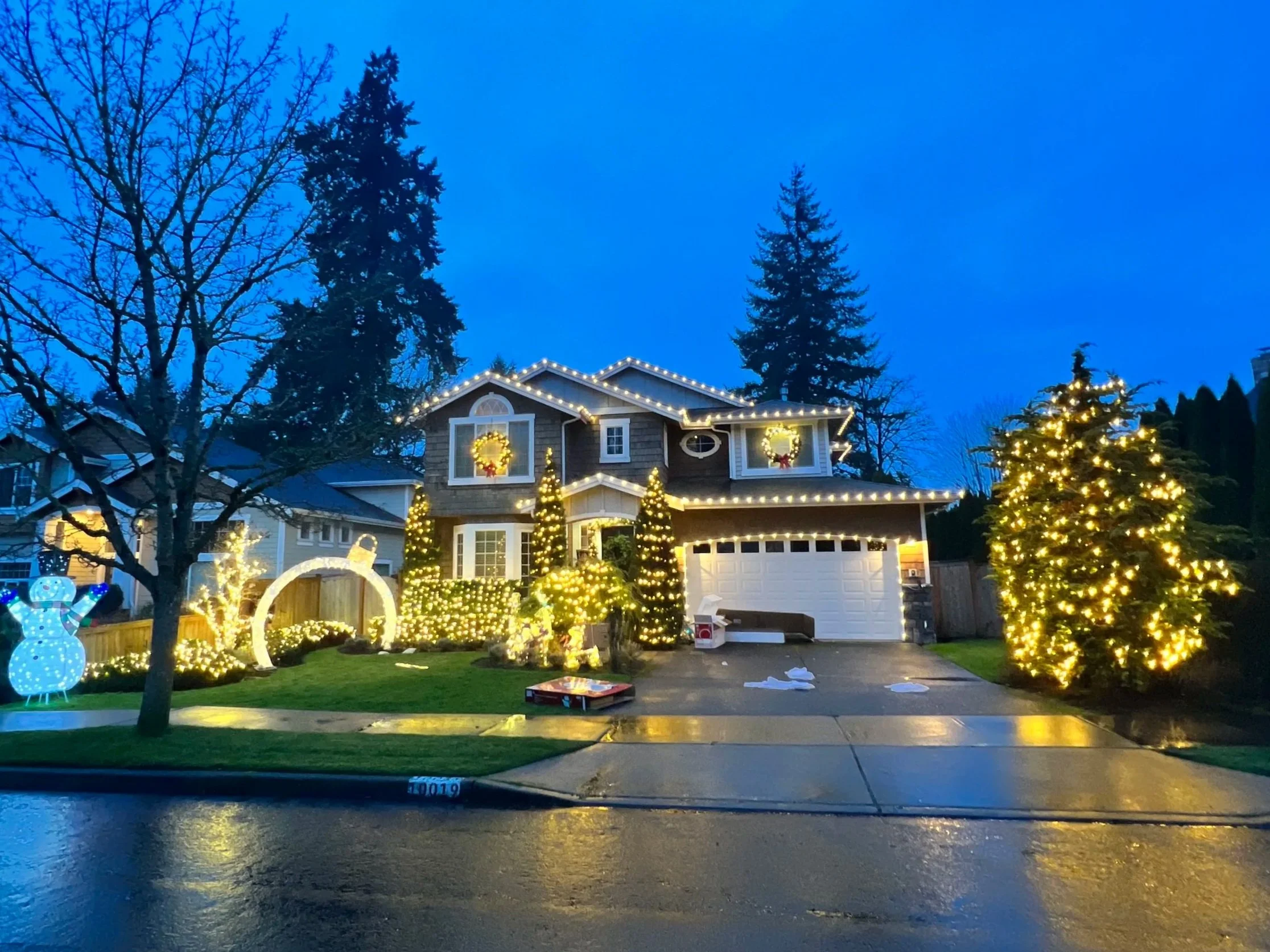 A house decorated with Christmas lights, wreaths, and illuminated trees at dusk, with a wet driveway reflecting the lights.