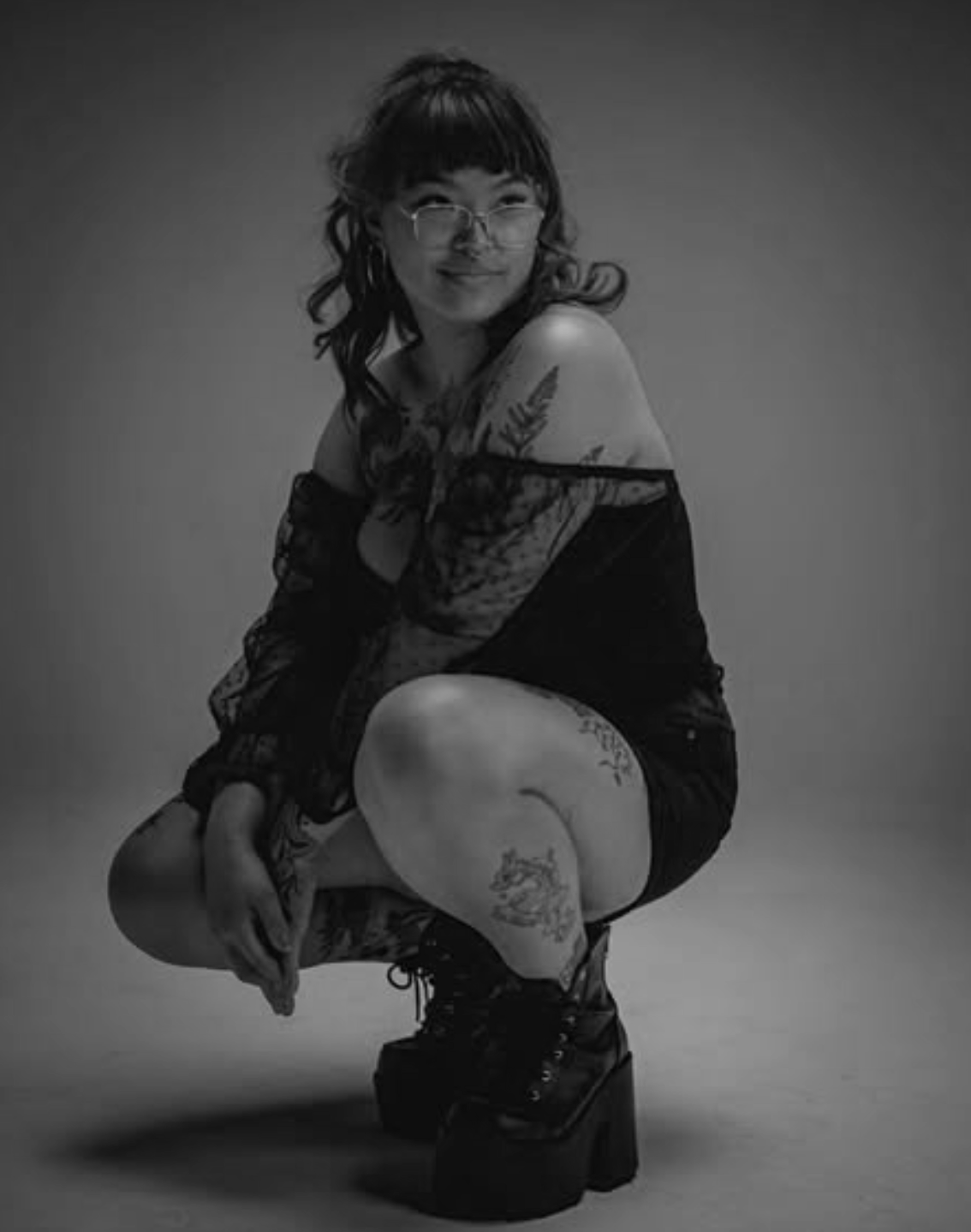 A person with glasses and tattoos squatting in platform boots, looking at the camera, black and white photo