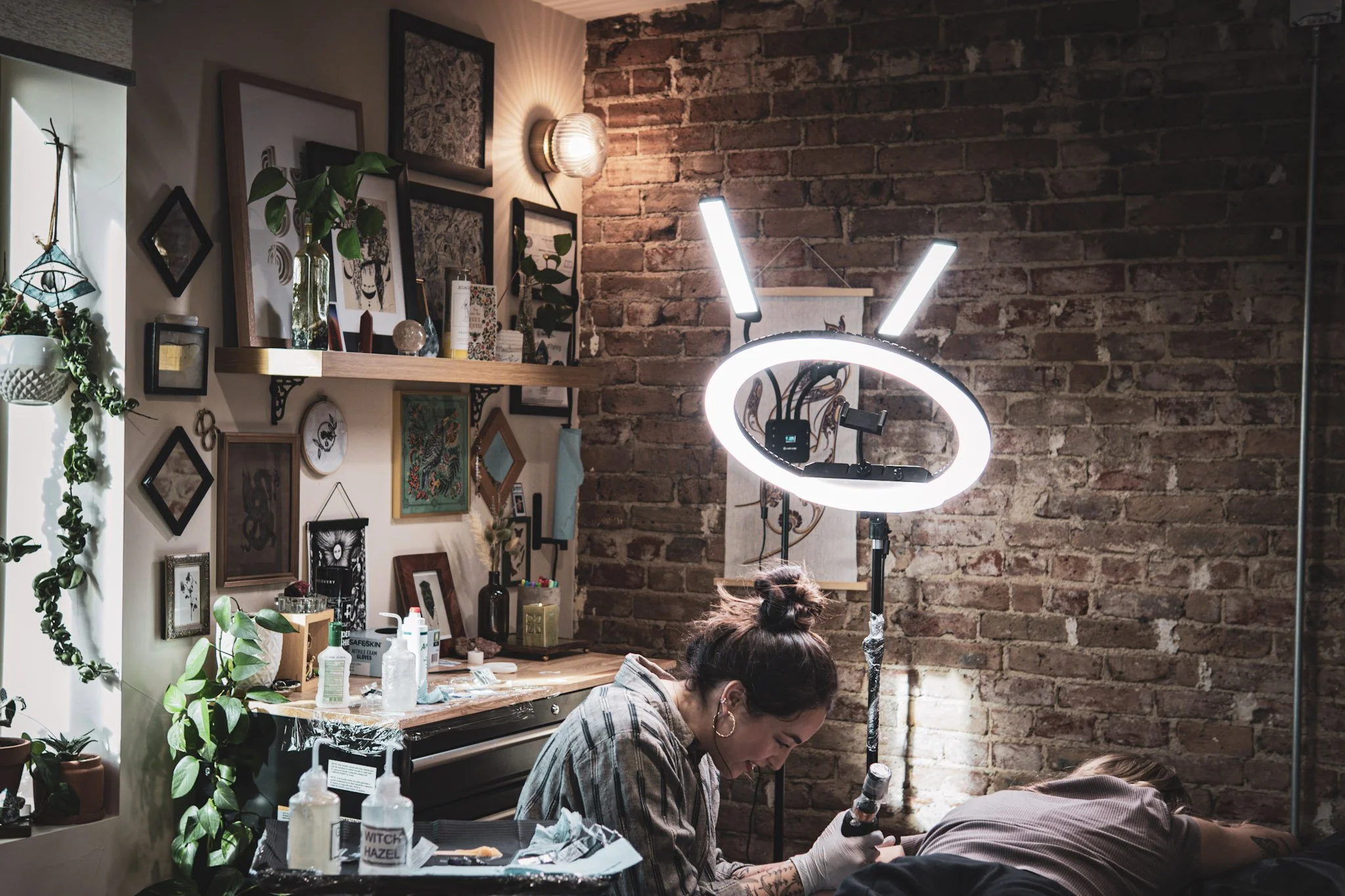 Tattoo artist working on a client in a studio with brick wall, gallery wall, plants, and ring light