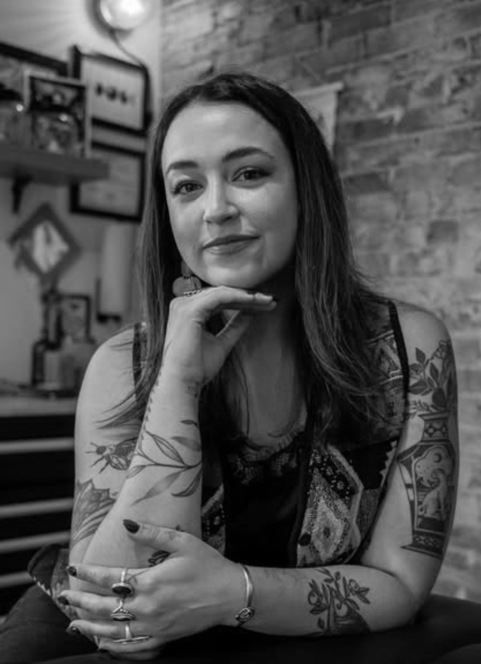 A woman with long dark hair and tattoos on her arms, sitting indoors with a brick wall background, looking at the camera.
