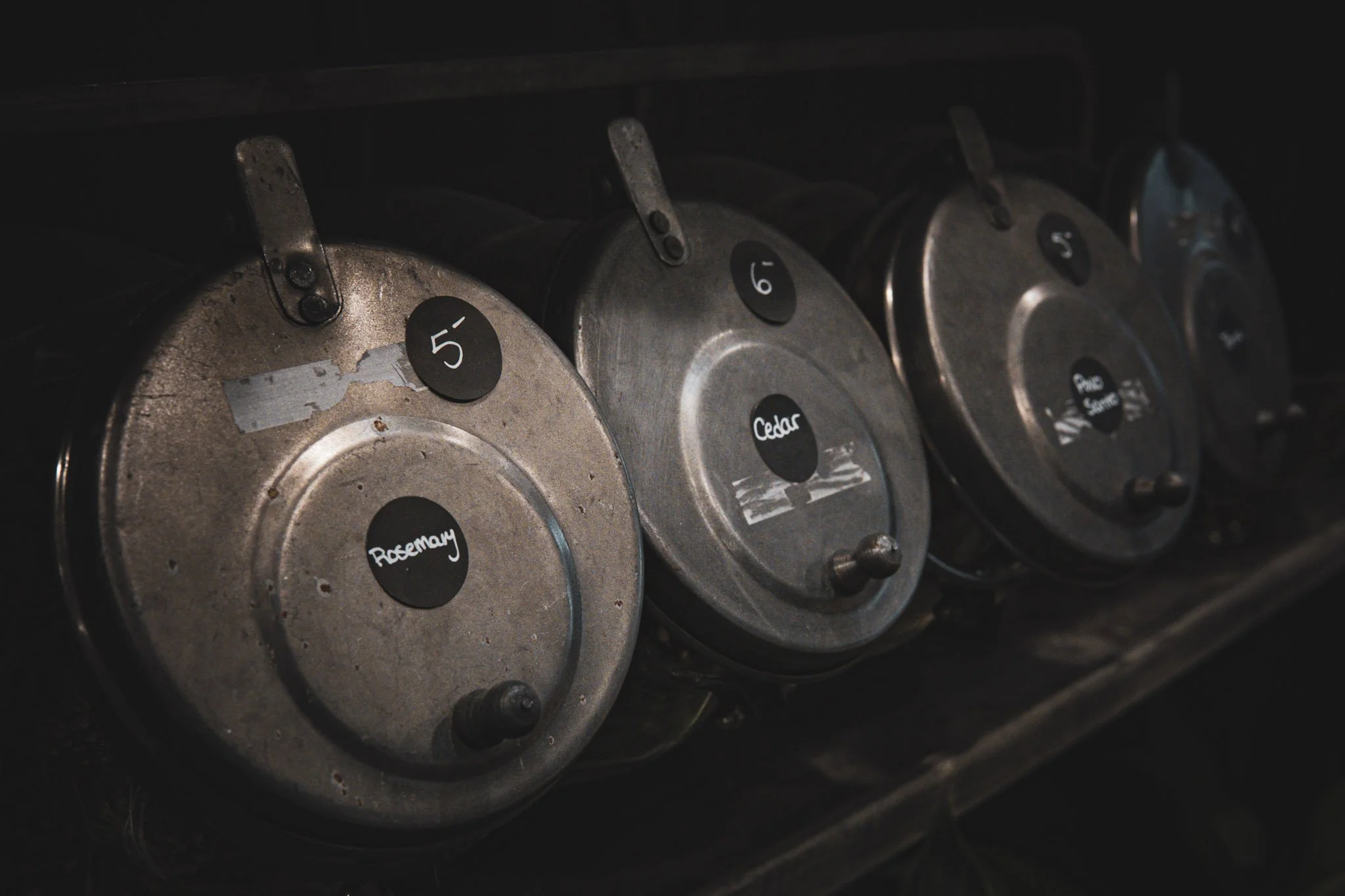 Metal cans with labels for rosemary, cedar, and pinesap, numbered 5, 6, and 7, stored on a dark shelf.
