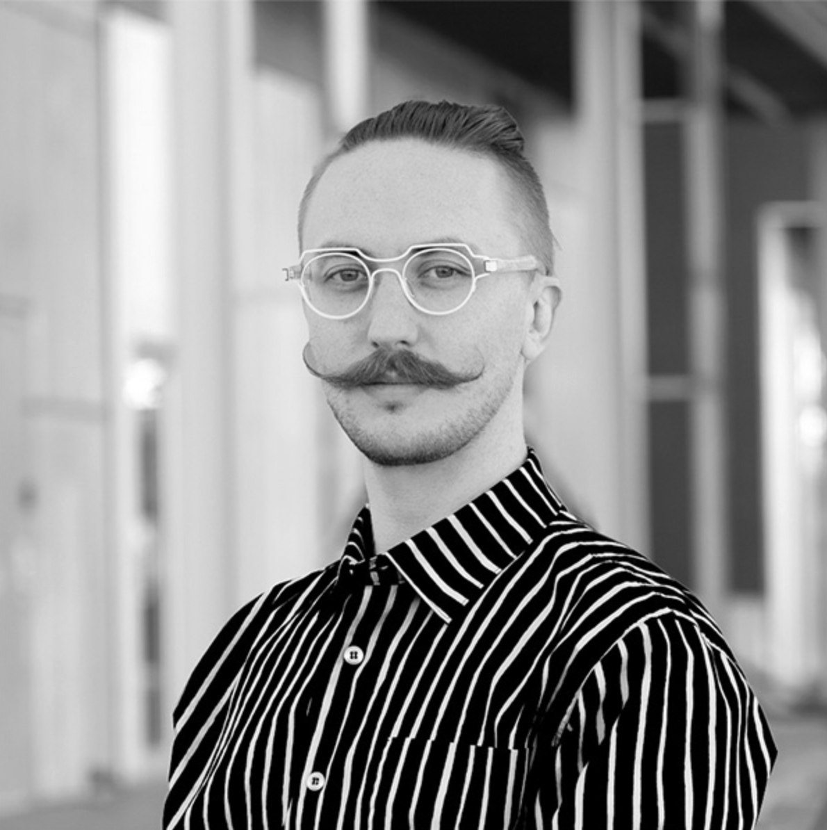Black and white portrait of a man with a styled mustache and beard, wearing round glasses and a striped shirt, standing in a modern indoor setting.